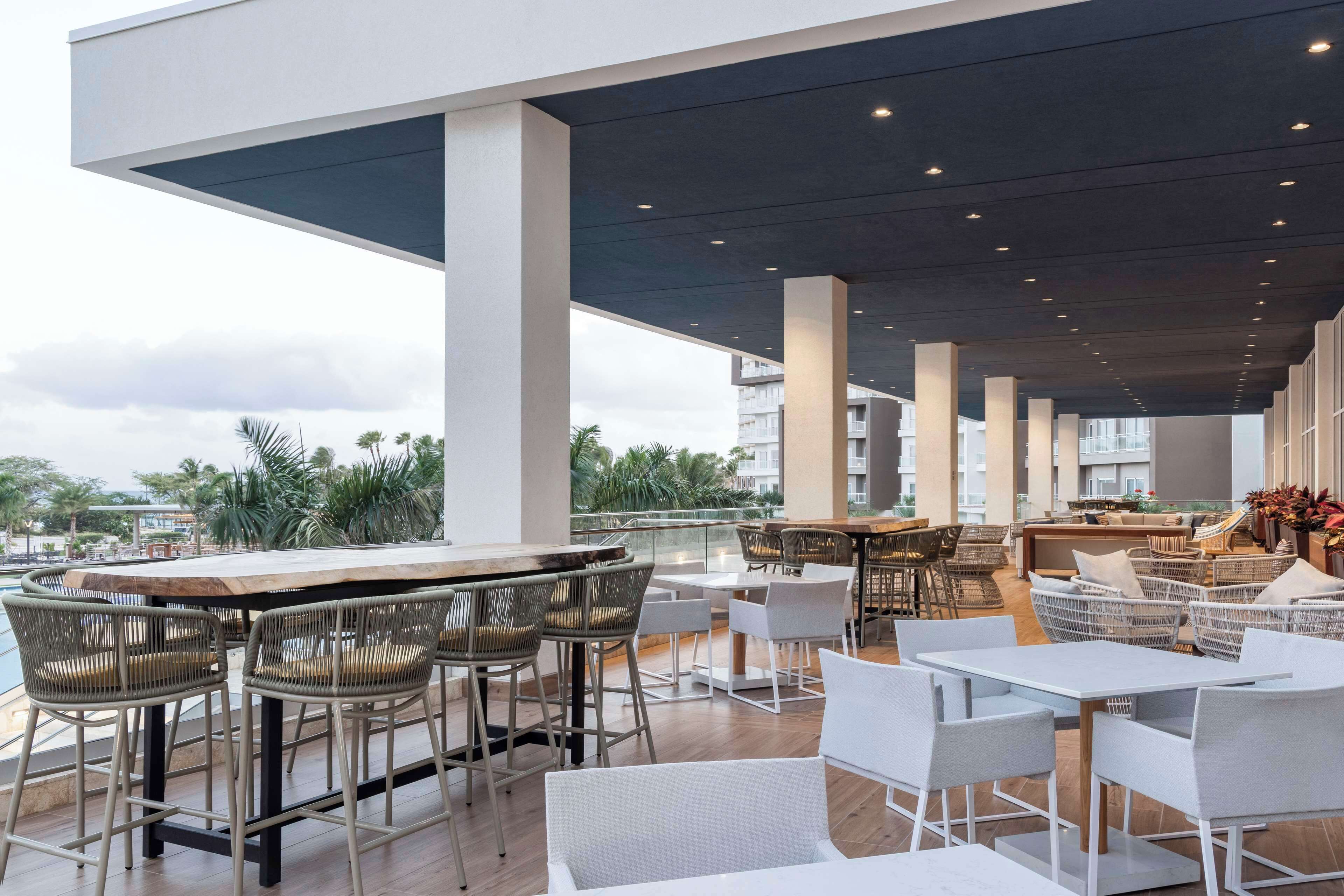 A rooftop bar with high tables and chairs, a pool, and a view of palm trees and buildings.