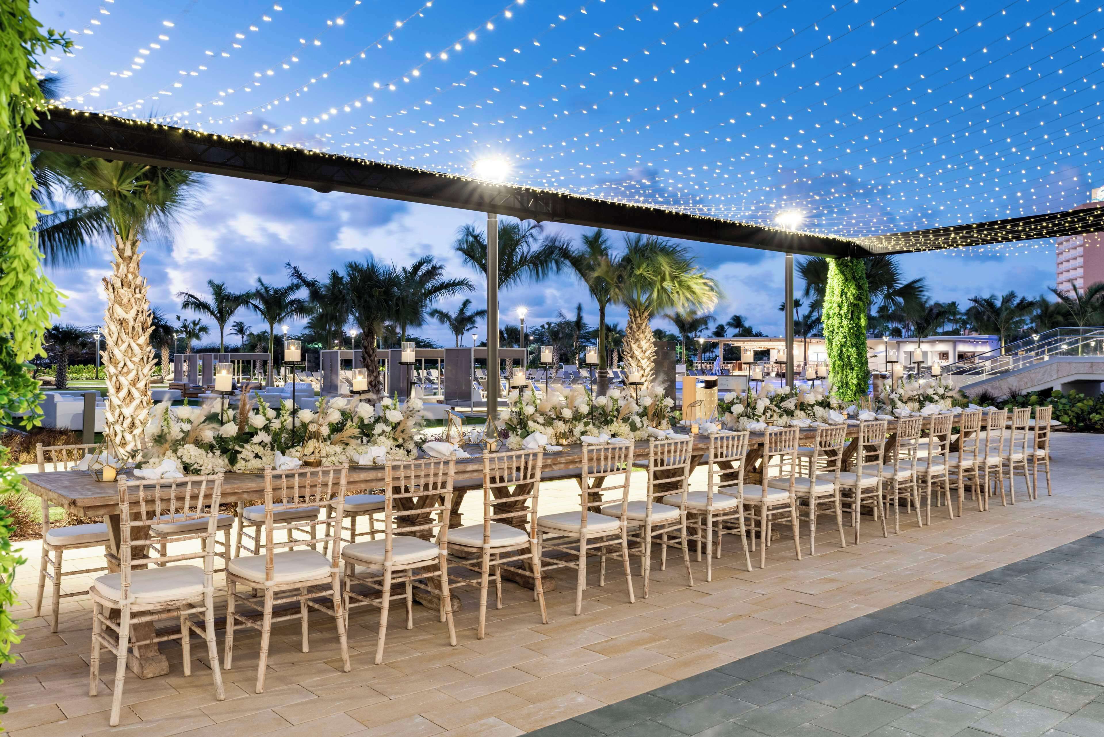 Un área de comedor al aire libre con varias mesas y sillas, decorada con flores y guirnaldas de luces.
