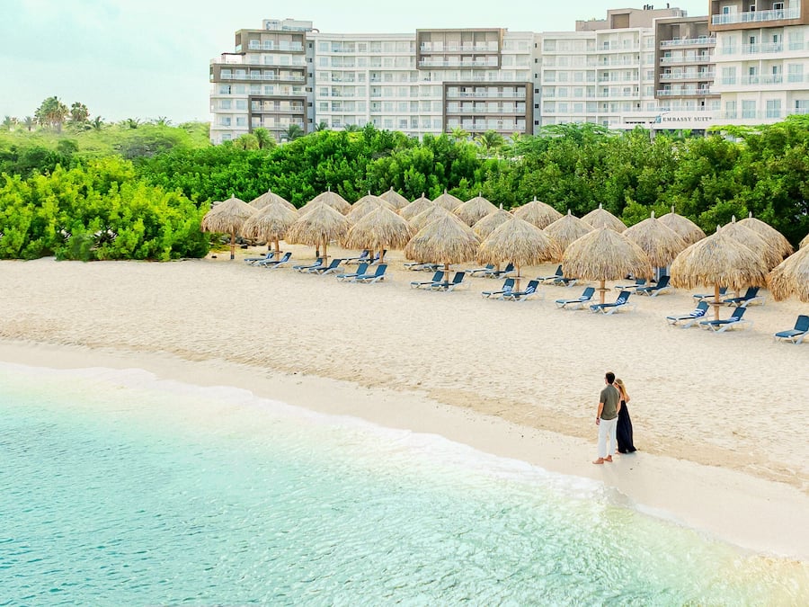 Una playa con filas de tumbonas y sombrillas de paja, mar azul transparente y un hotel al fondo.