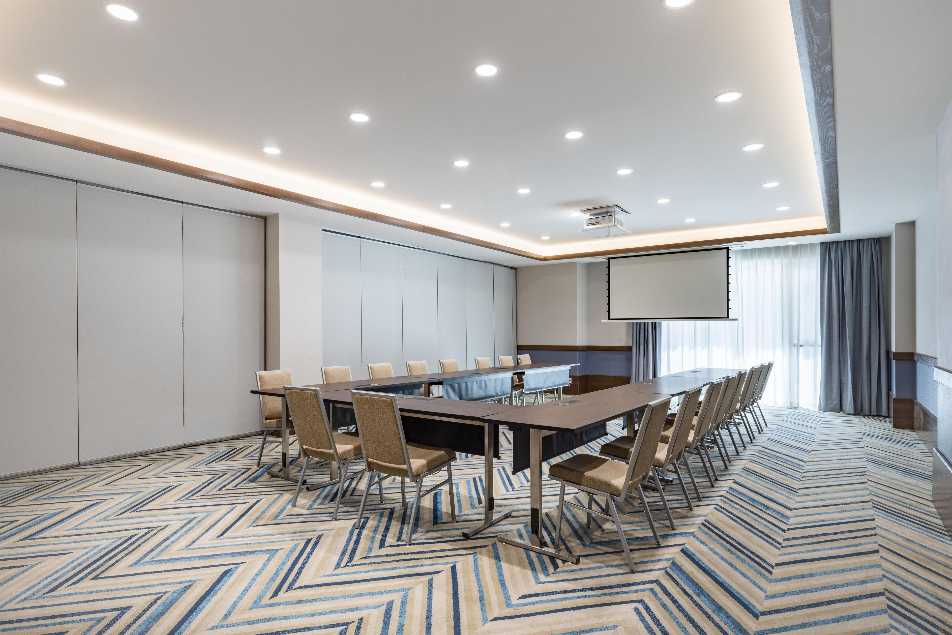 Un salón de conferencias con una larga mesa, sillas, una pantalla de proyector y un amplio ventanal con cortinas.