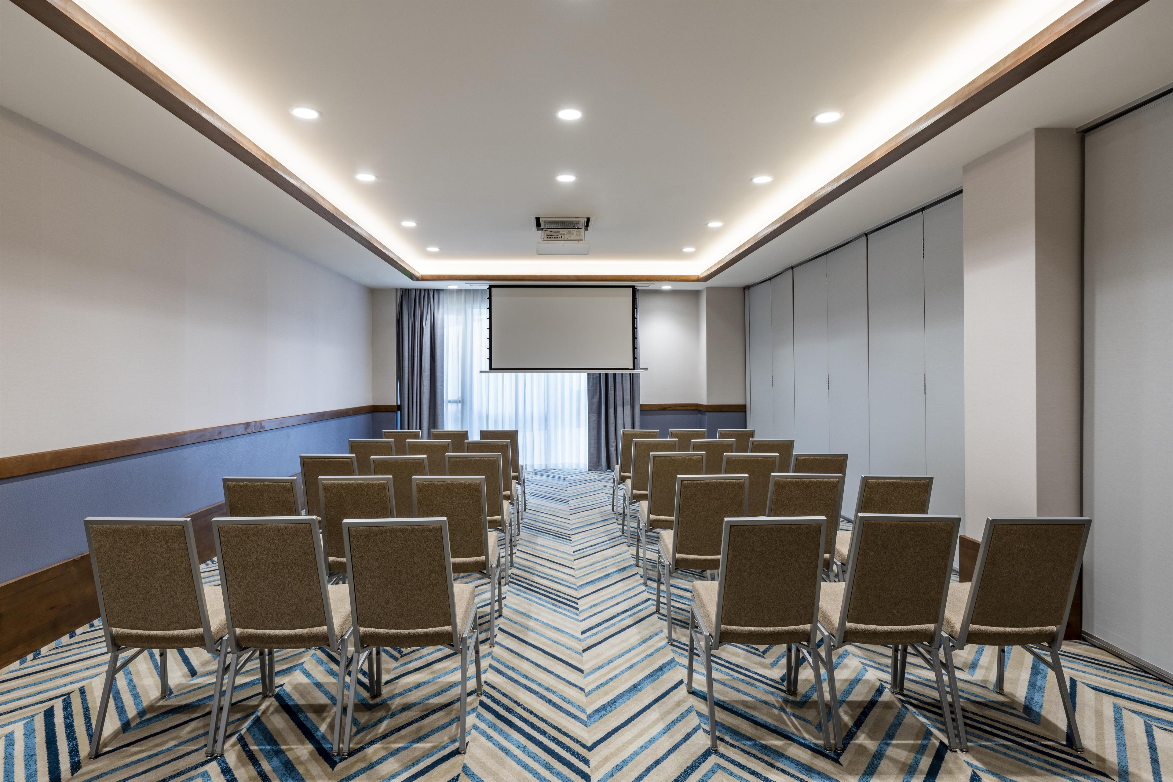 A conference room with rows of chairs, a projector screen, and a carpeted floor.