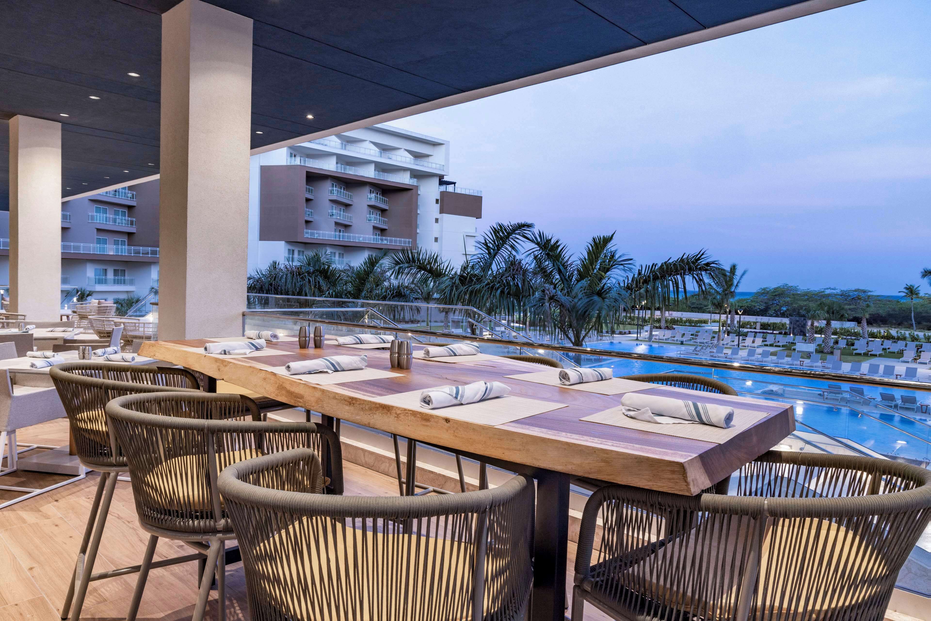 Zona de comedor junto a la piscina con sillas de mimbre y una mesa de madera, con vistas a la piscina y un edificio con balcones.