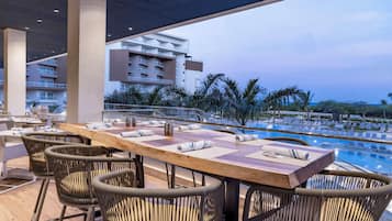 Zona de comedor junto a la piscina con sillas de mimbre y una mesa de madera, con vistas a la piscina y un edificio con balcones.