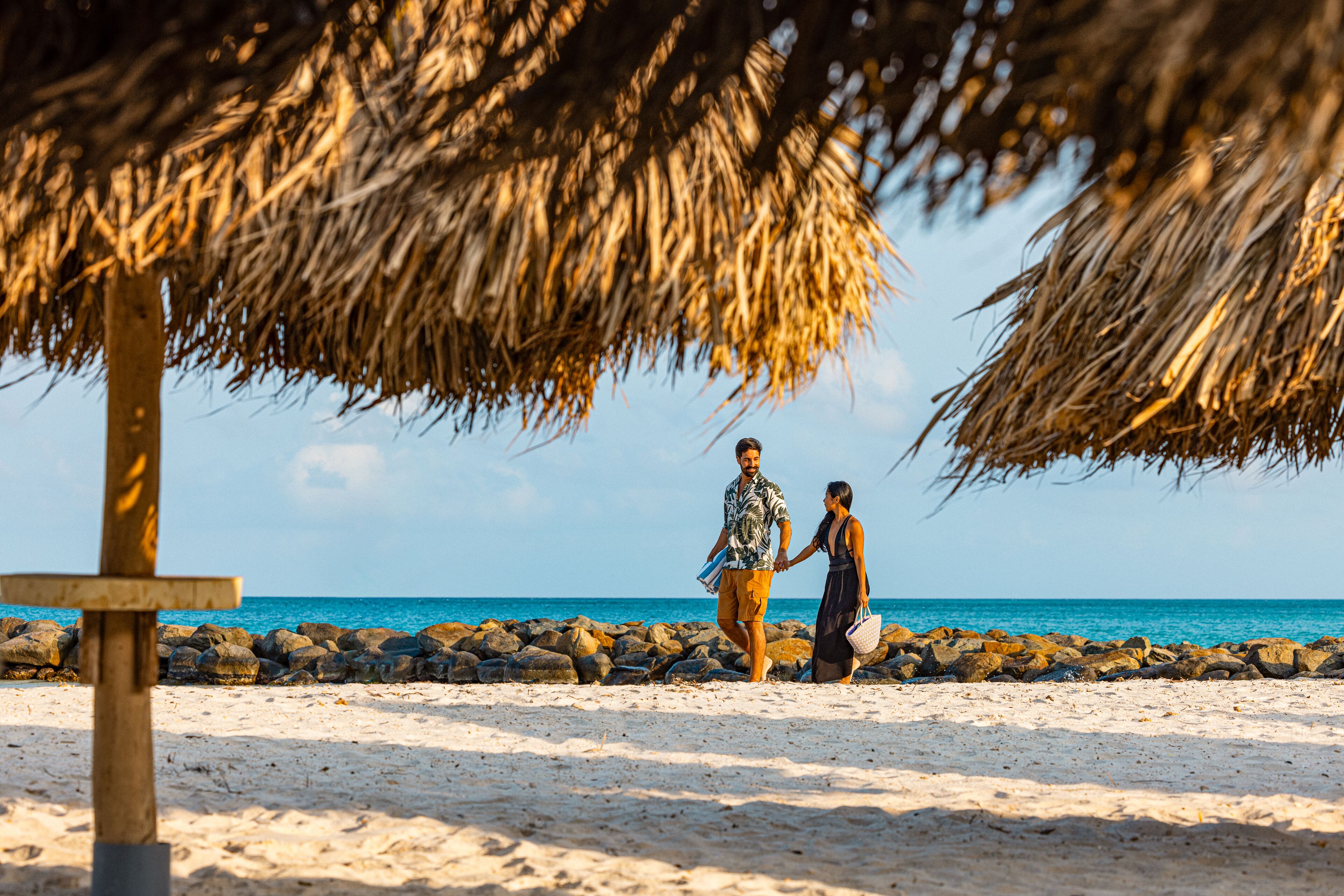A couple walking on a sandy beach with a thatched umbrella and a wooden signpost.