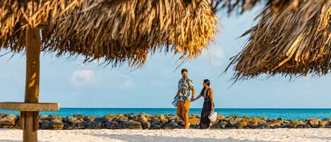 A couple walking on a sandy beach with a thatched umbrella and a wooden signpost.
