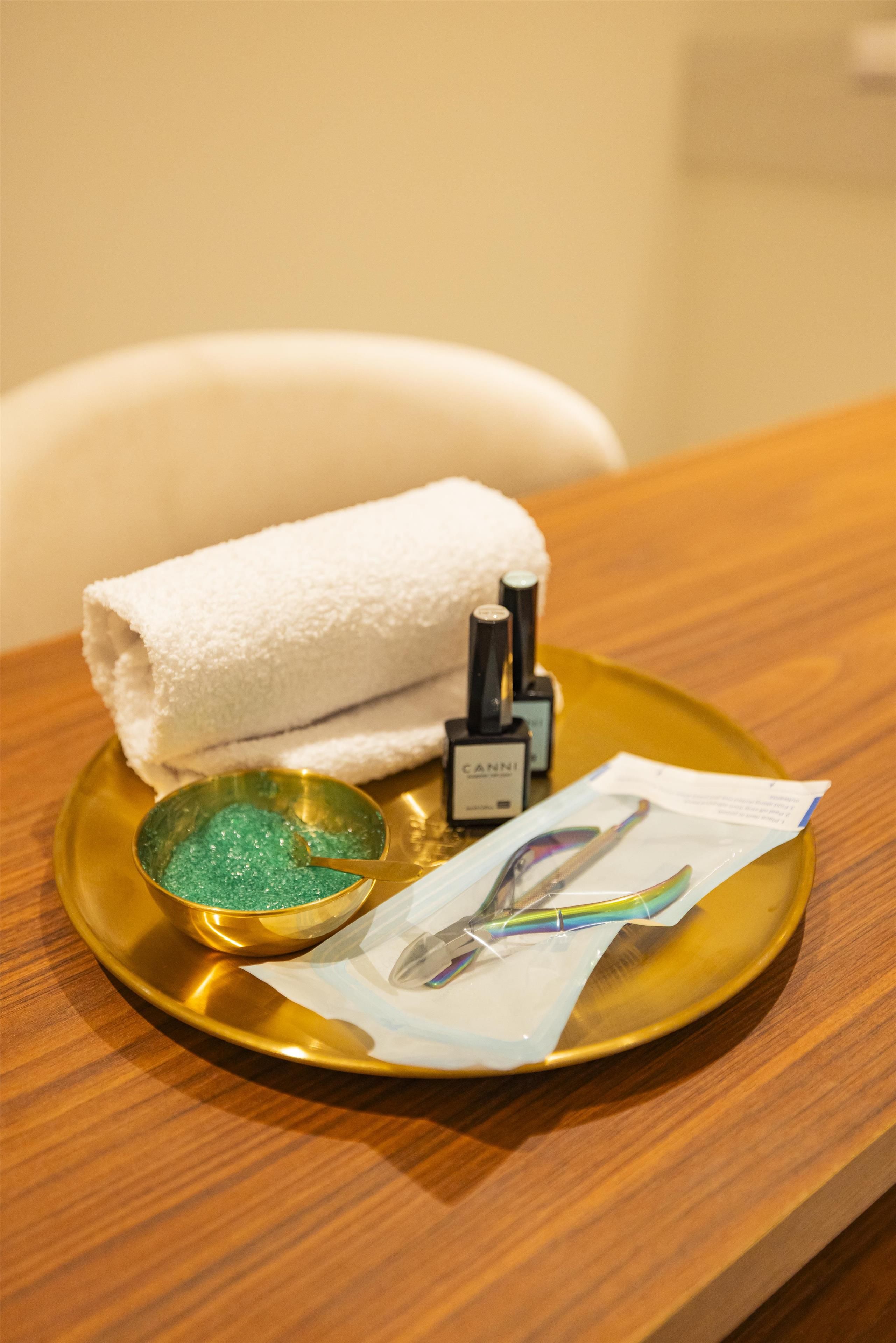 A tray with a white towel, a bottle of Canni body oil, a bowl of green bath salts, and a pair of sunglasses on a wooden table.