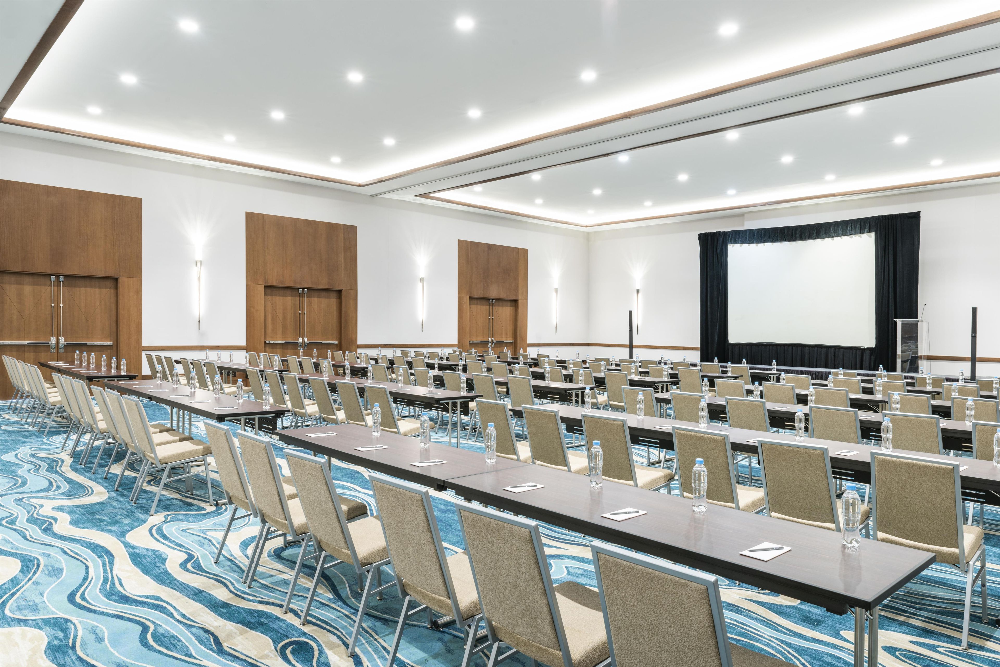 Un salón de conferencias con filas de mesas y sillas, una pantalla grande y un escenario.