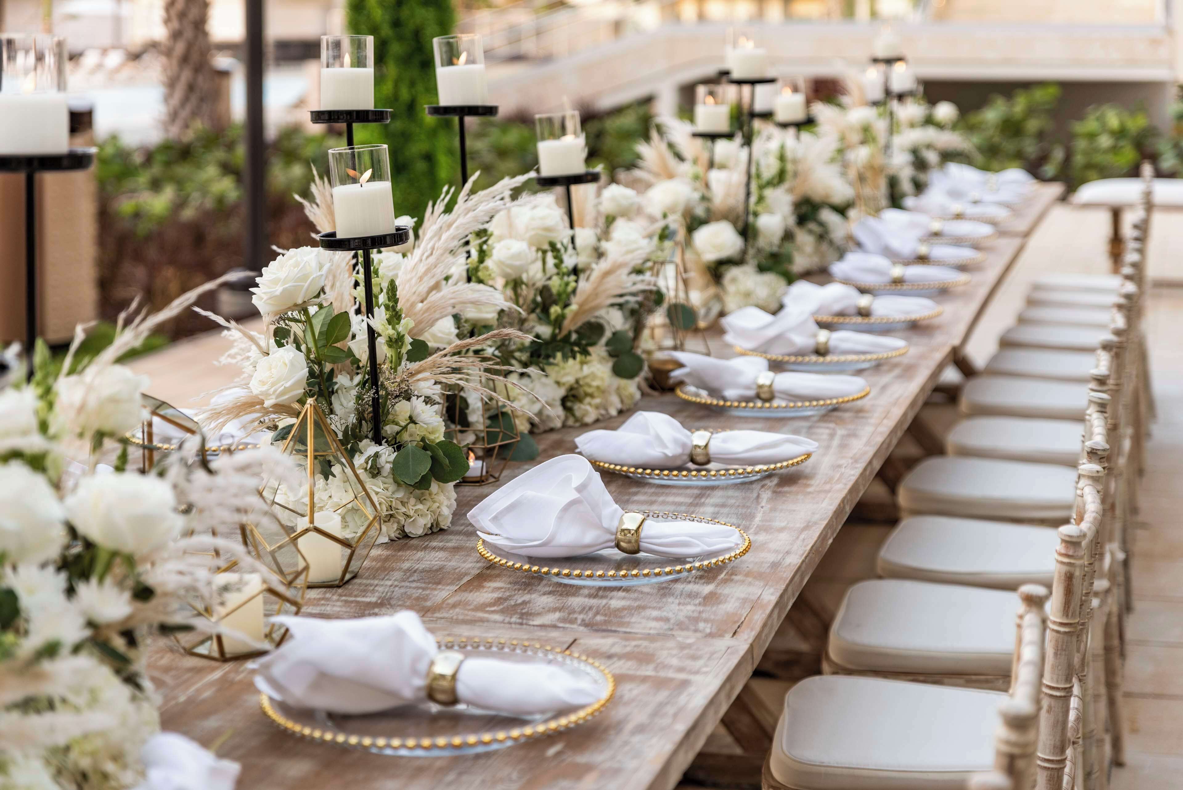 A long wooden table set with white napkins, gold beaded napkin rings, and white plates, surrounded by white flowers and candles.