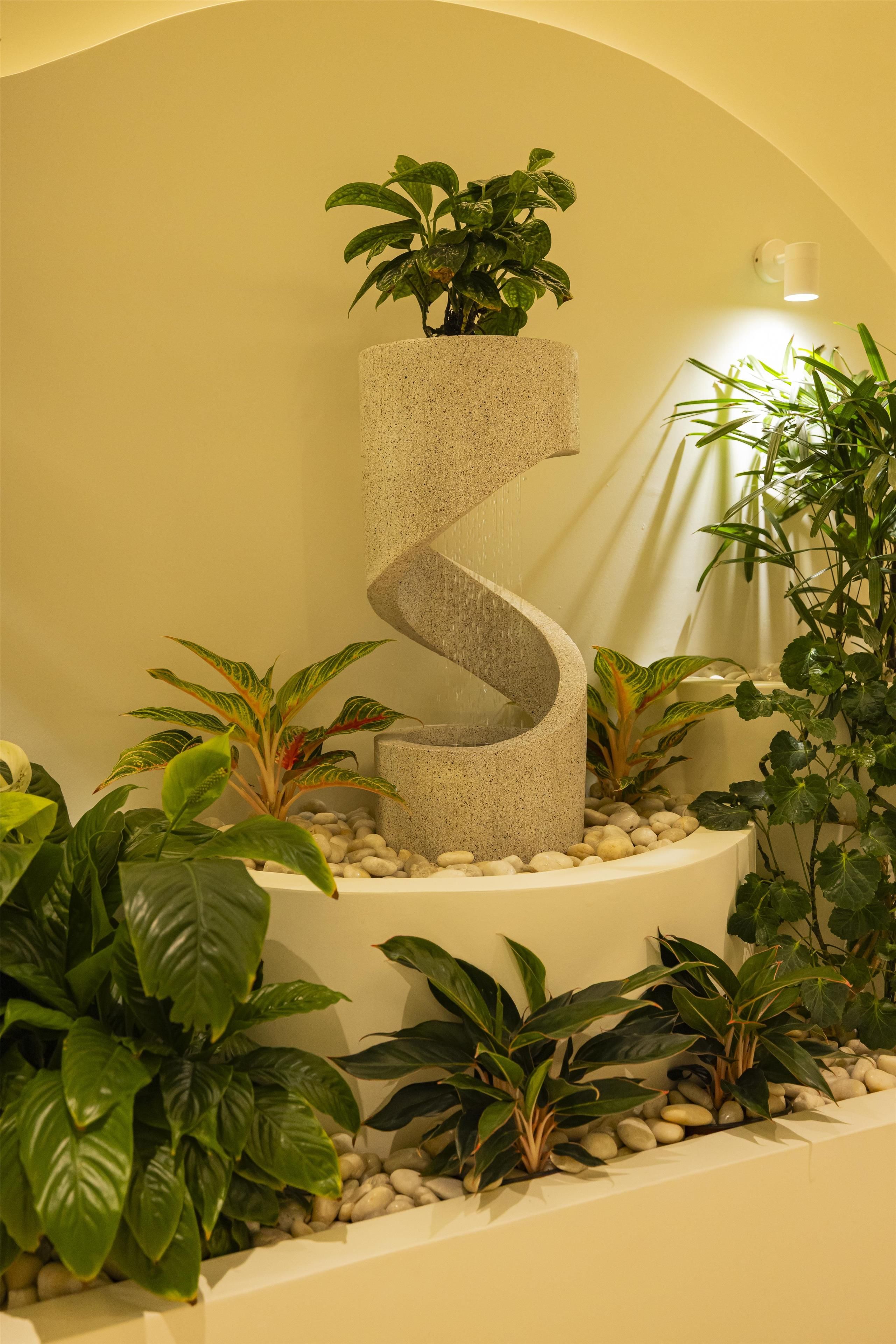 A spiral stone planter with a potted plant, surrounded by various other plants and pebbles.