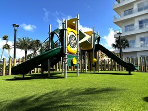 Un parque infantil con toboganes y estructuras para escalar, rodeado de césped y palmeras, con un edificio al fondo.