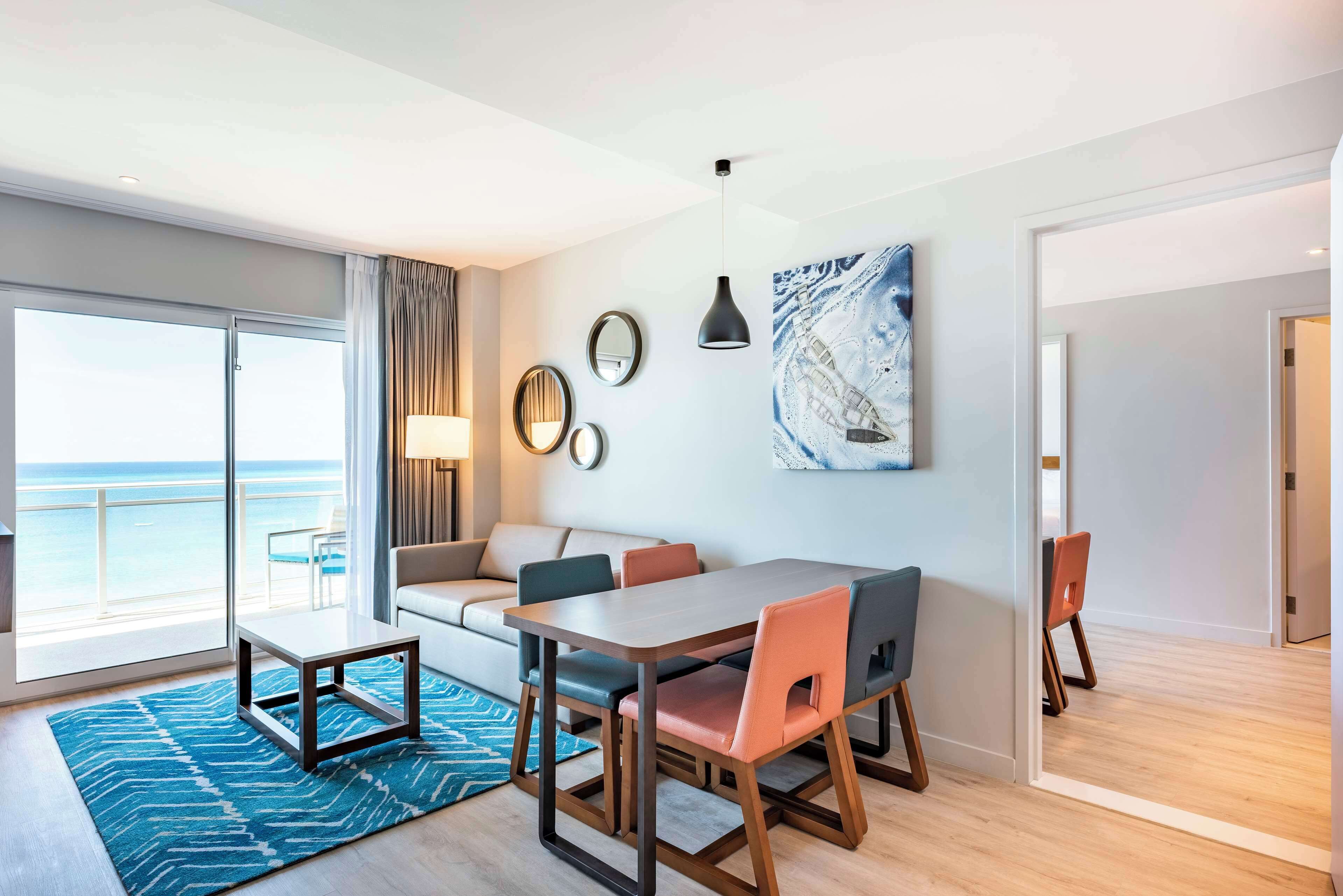 A modern hotel room with a balcony, a dining area, and a large window offering a view of the sea.