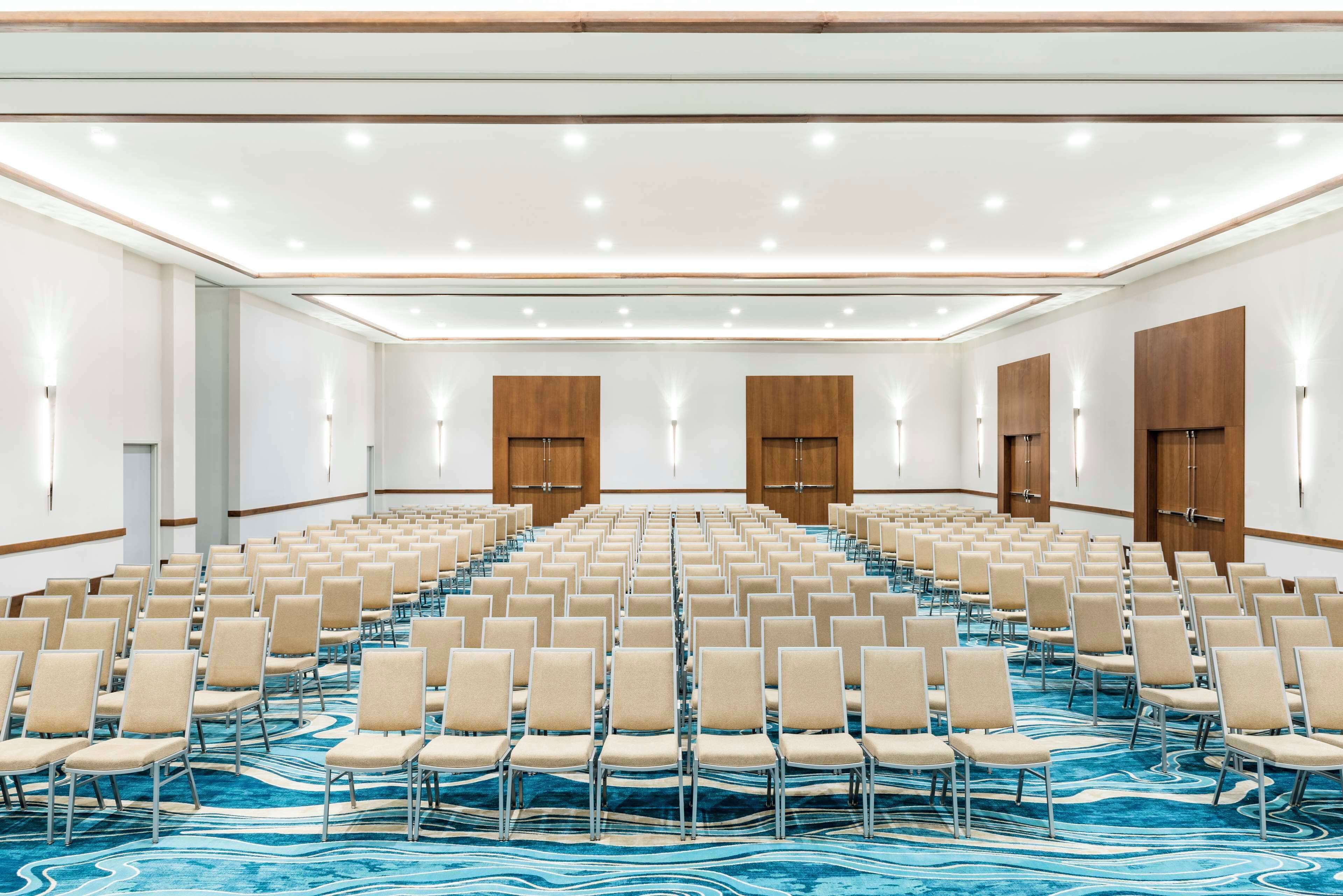 Un amplio salón de conferencias con filas de sillas, puertas de madera y una alfombra azul estampada.
