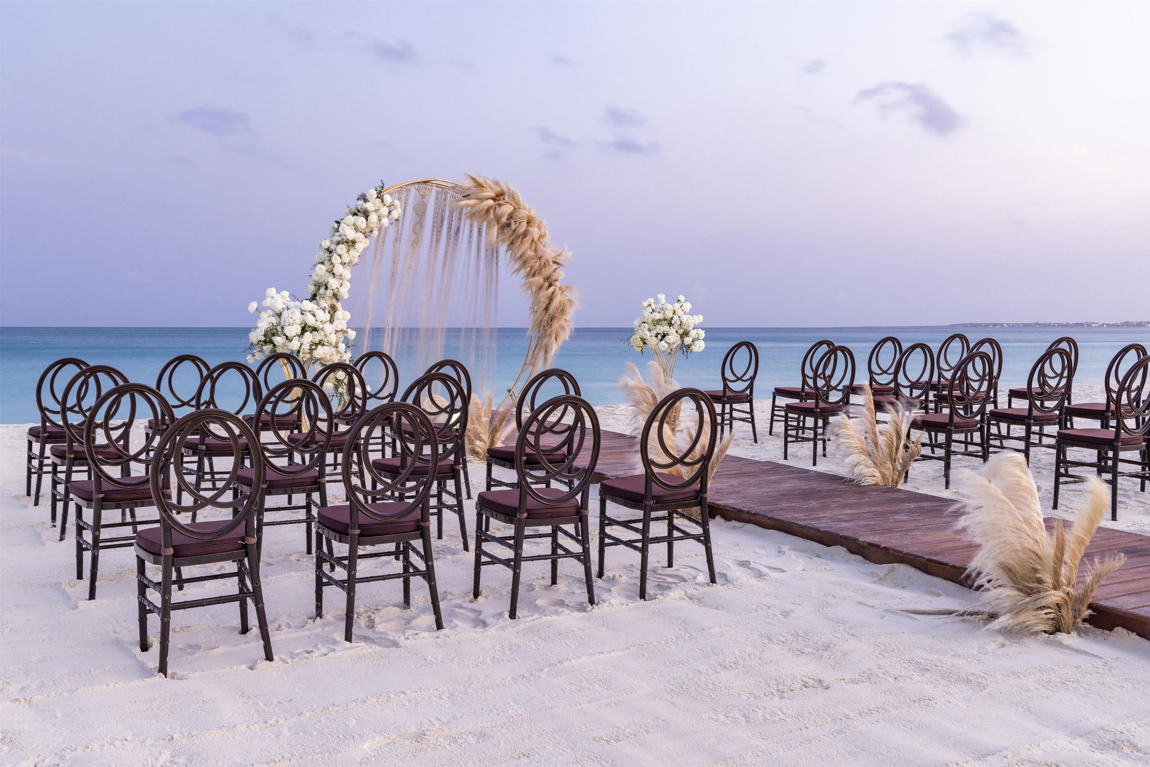 A beach wedding setup with a wooden aisle, chairs, and floral decorations.