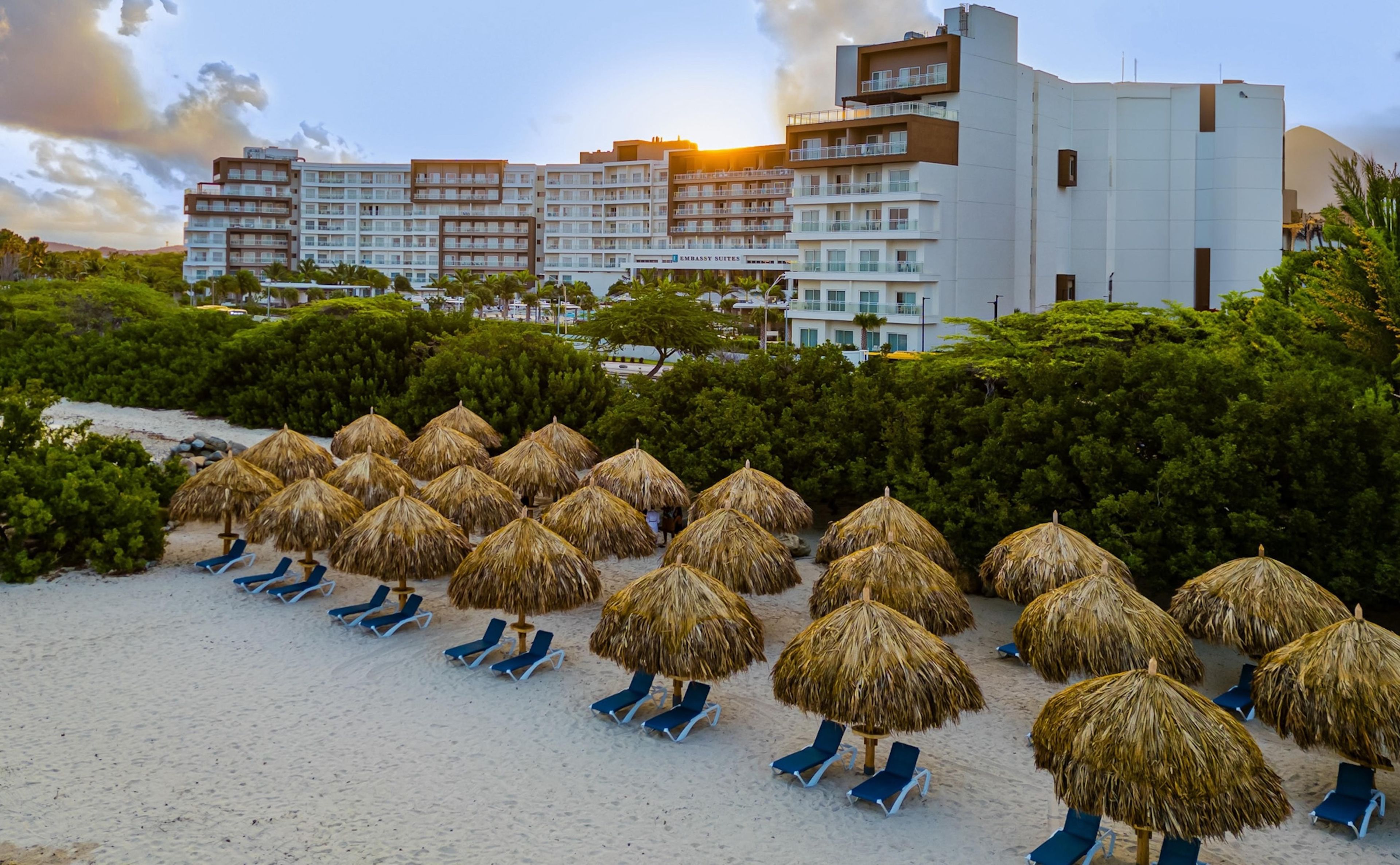Ett hotell vid stranden med rader av parasoller med halmtak och blå solstolar.