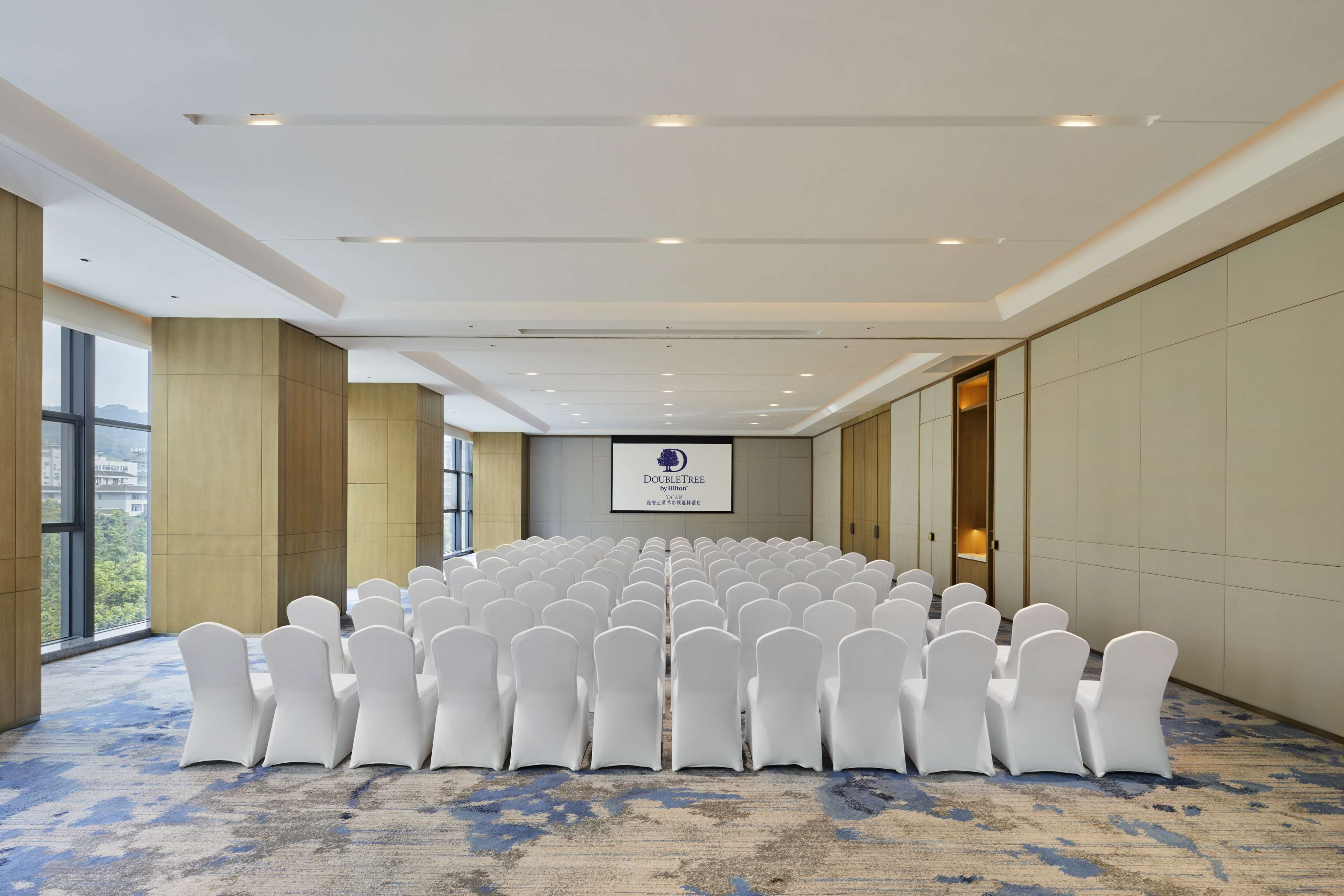 A conference room with rows of white chairs, a large screen, and a view of the outdoors.