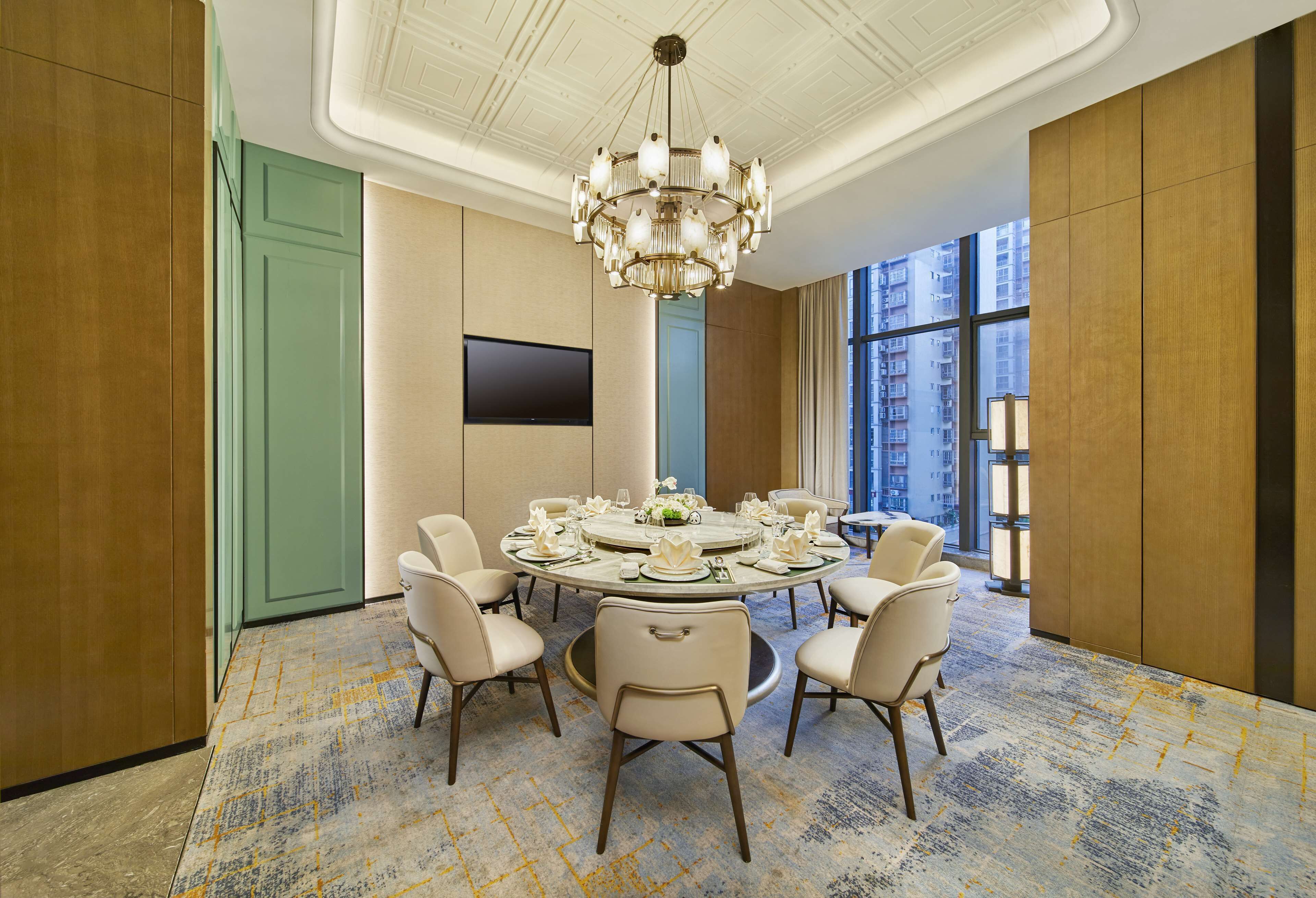 A conference room with a round table, six chairs, a chandelier, a flat-screen TV, and large windows with a city view.