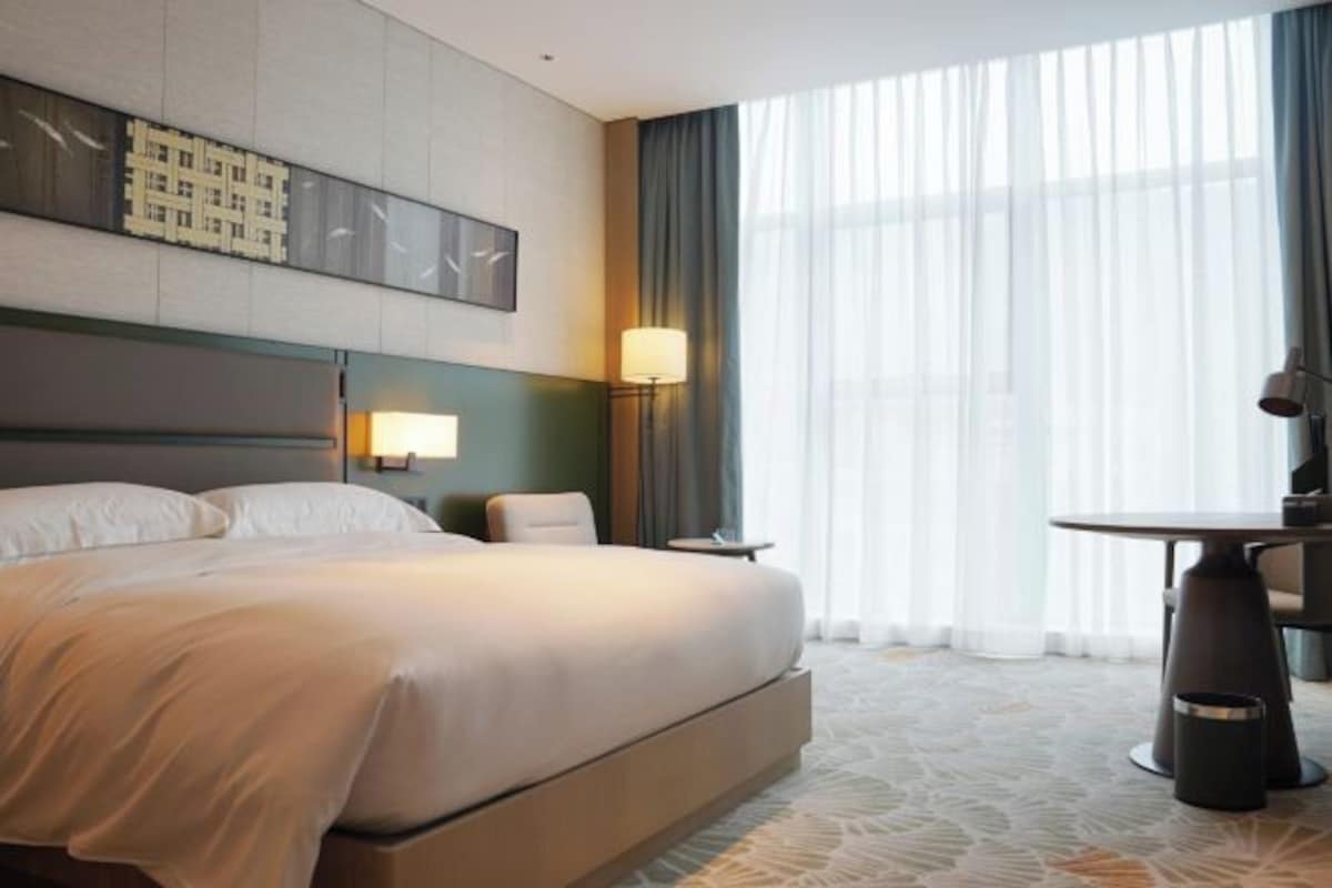 A hotel room with a large bed, a desk, and a chair. There is a lamp on the desk and a window with curtains.