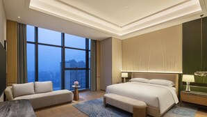 Una habitación de hotel moderna con una cama grande, un sofá, una mesita auxiliar y vistas a la ciudad a través de grandes ventanales.