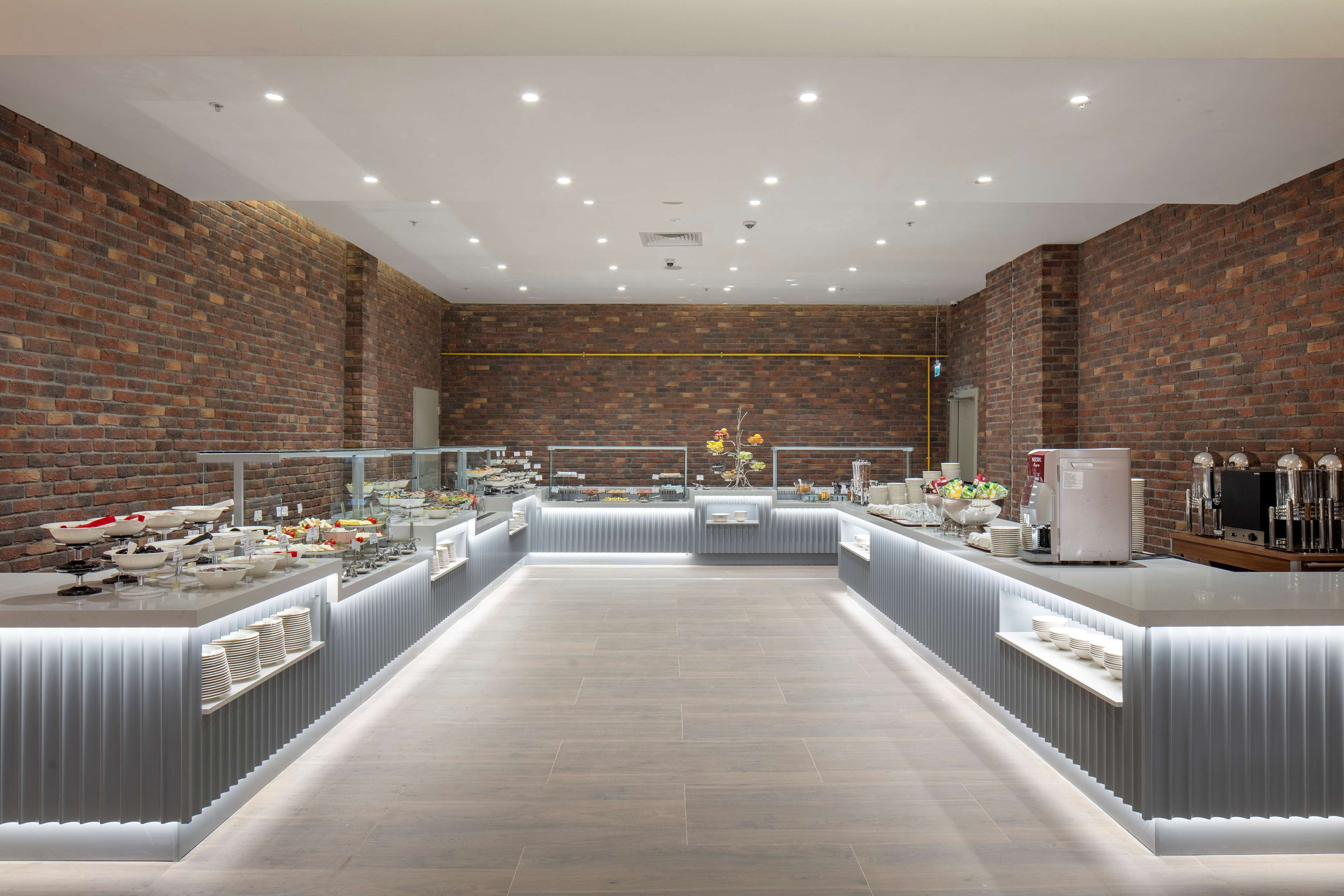 Een moderne ontbijtbalk met een achtergrond van een bakstenen muur, een lange toonbank met diverse etenswaren en een nette betegelde vloer.
