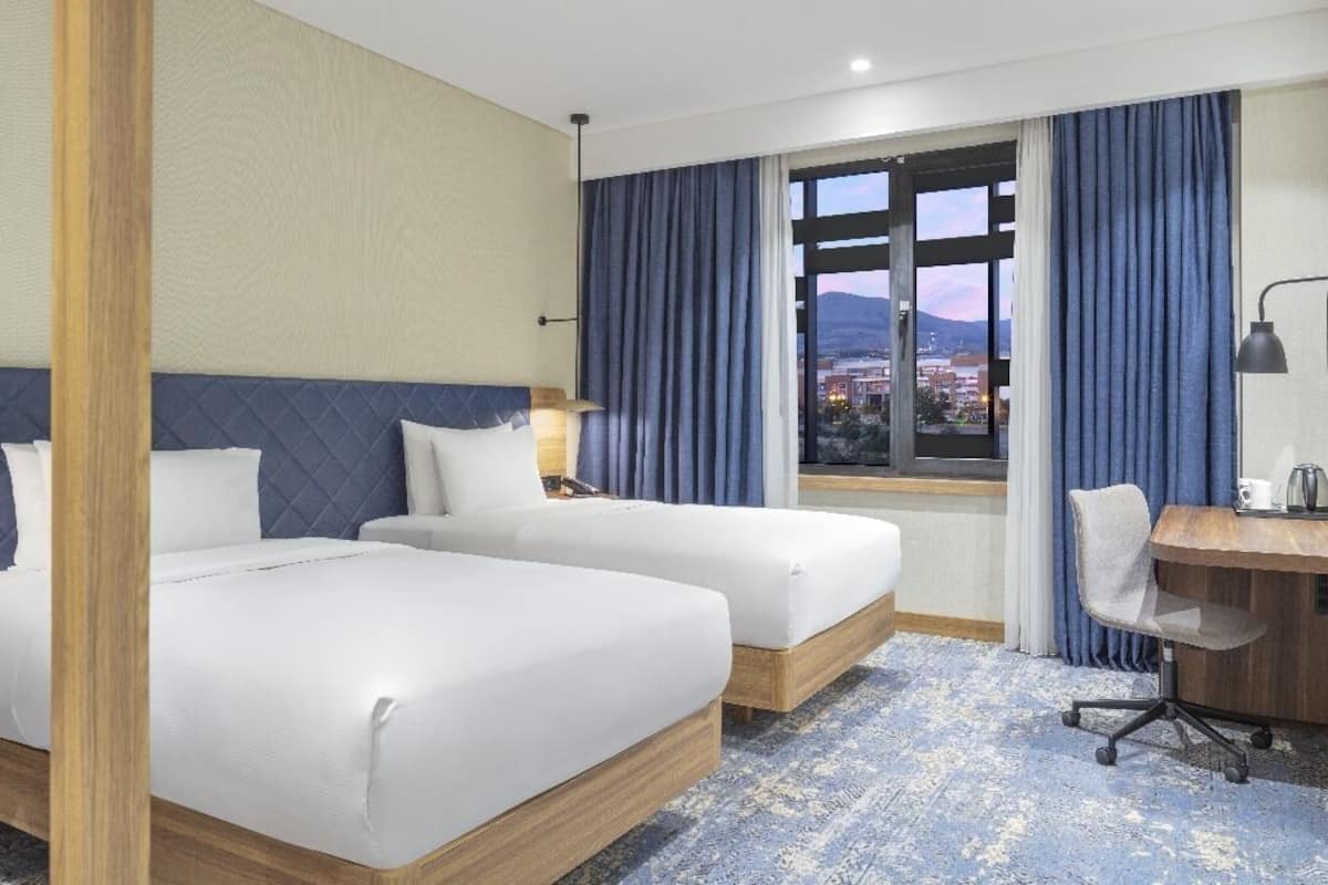 Ein Hotelzimmer mit zwei Betten, einem Schreibtisch, einem Stuhl, einem großen Fenster mit Vorhängen und Blick auf die Stadtlandschaft.