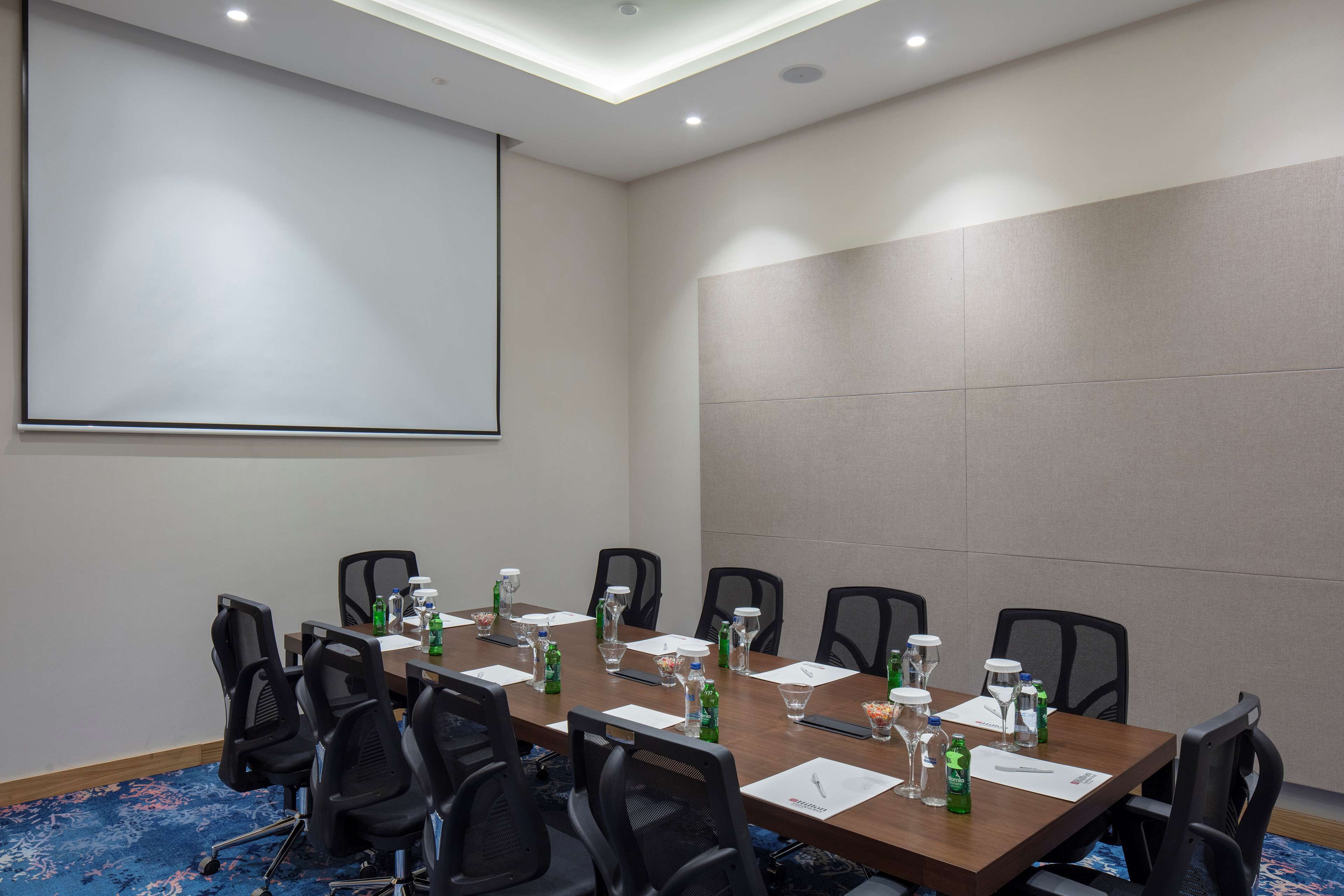A conference room with a long table, chairs, a projector screen, and various items like water bottles and papers.