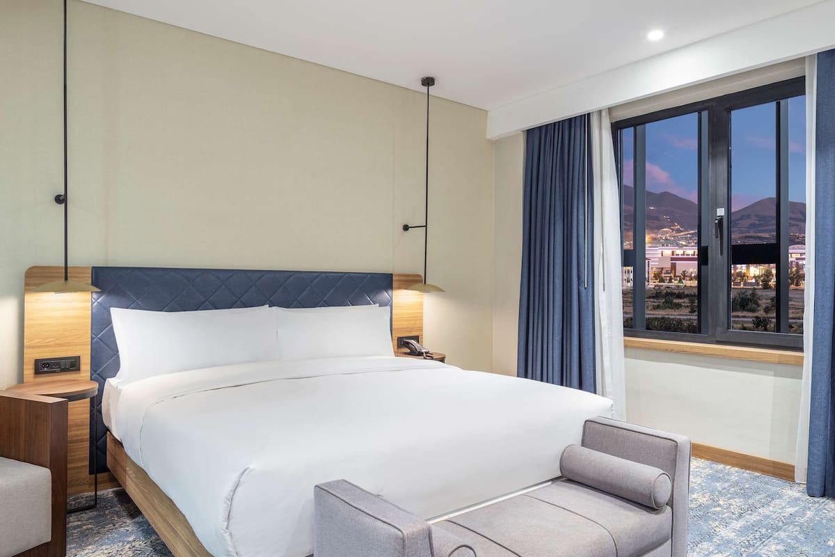 Ein Hotelzimmer mit Bett, Schreibtisch, Stuhl, Nachttisch und Fenster mit Blick auf eine Stadtlandschaft.
