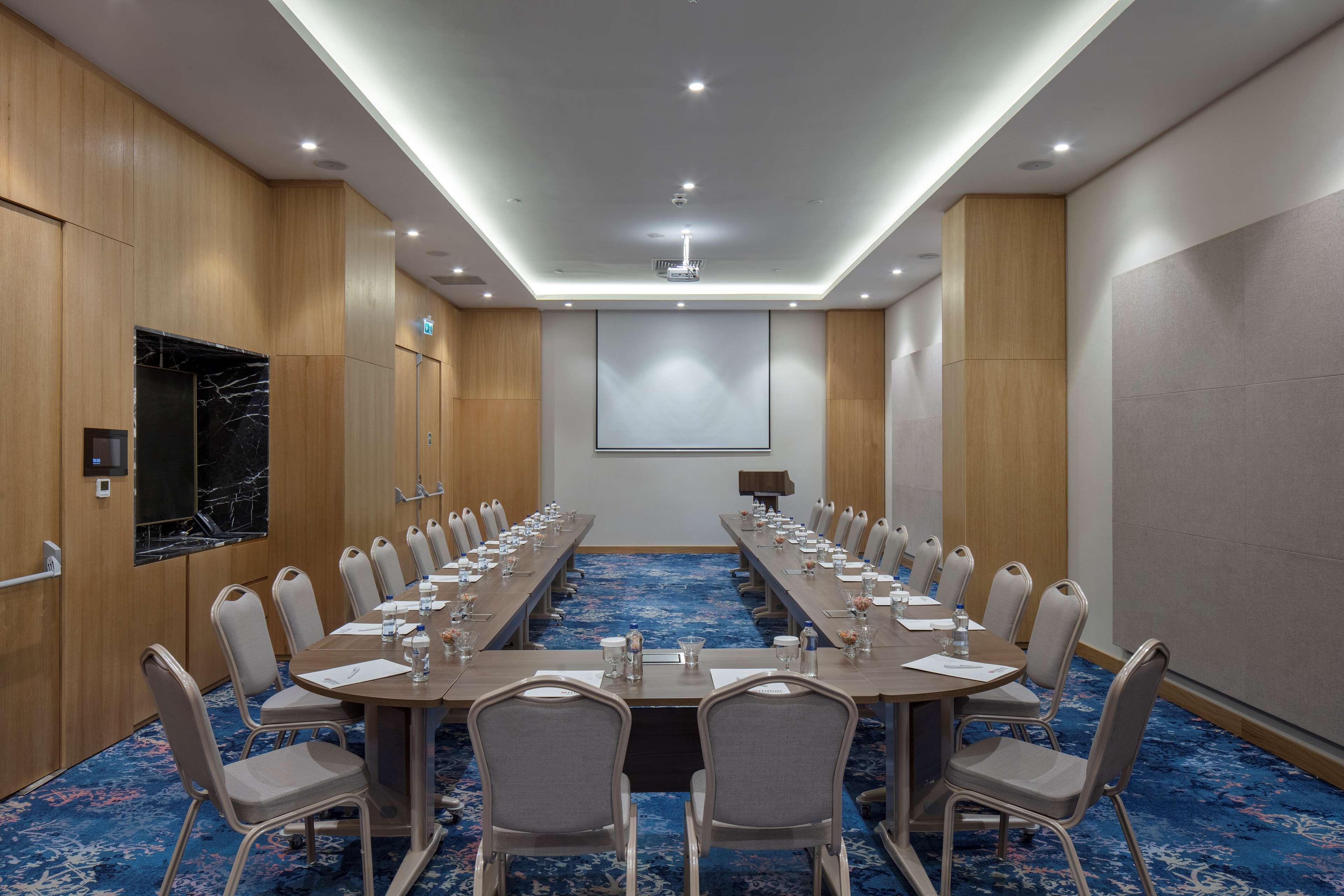 Ett konferensrum med ett långt bord, stolar, en projektorduk och en vägg som har en dekorativ marmorbeklädnad.