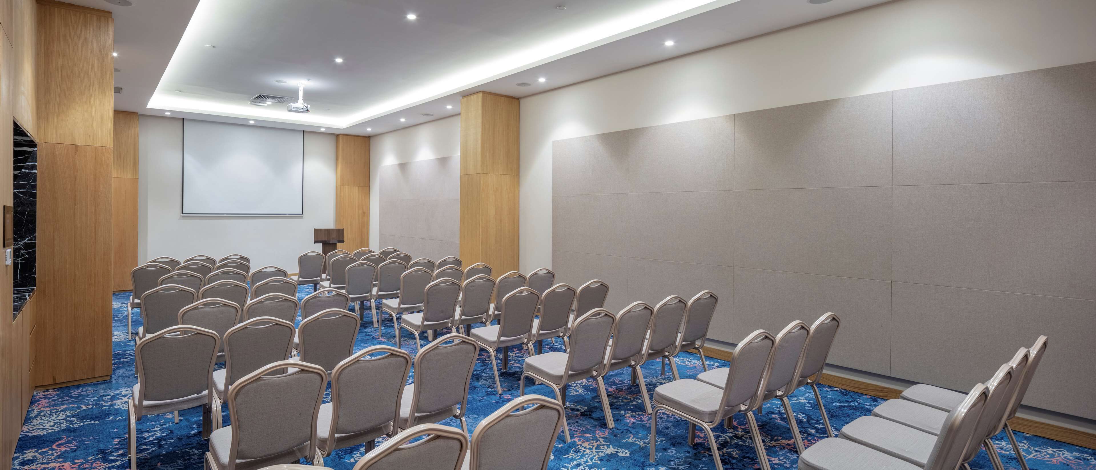 A conference room with rows of chairs, a projector screen, and a carpeted floor.