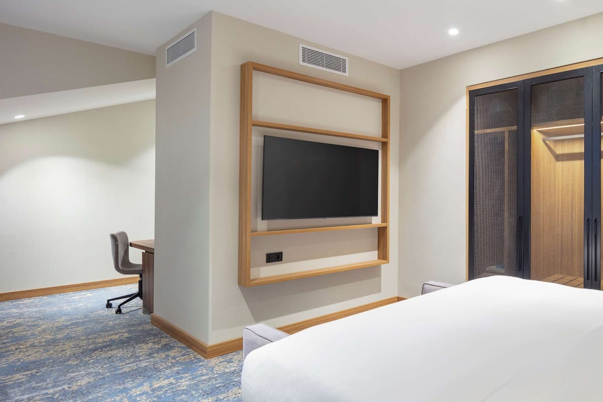 Ein Hotelzimmer mit einem Bett, einem an der Wand montierten Fernseher, einem Schreibtisch mit Stuhl und einem großen Fenster mit Jalousien.