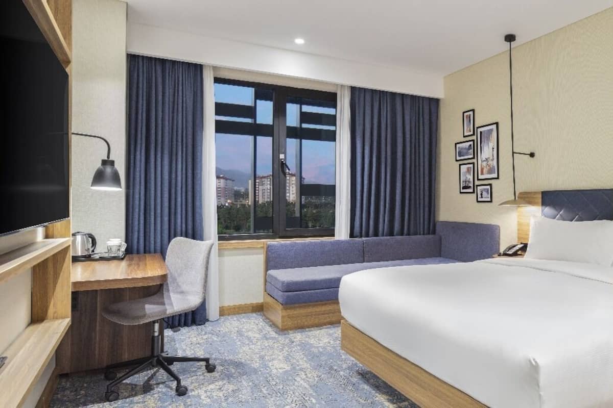 Ein Hotelzimmer mit Bett, Schreibtisch, Stuhl, Fernseher, einem Fenster mit Vorhängen und Blick auf die Stadt.