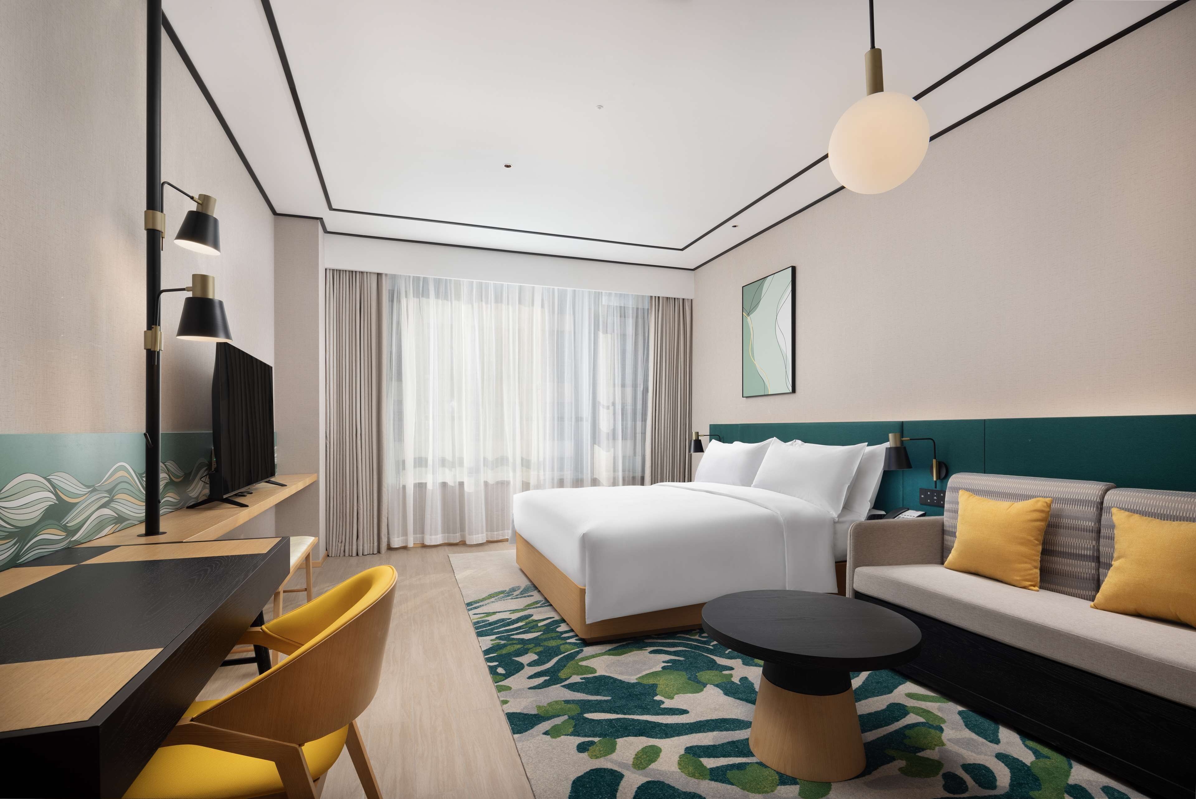 A modern hotel room with a large bed, a sofa, a desk with chairs, and a dining table.