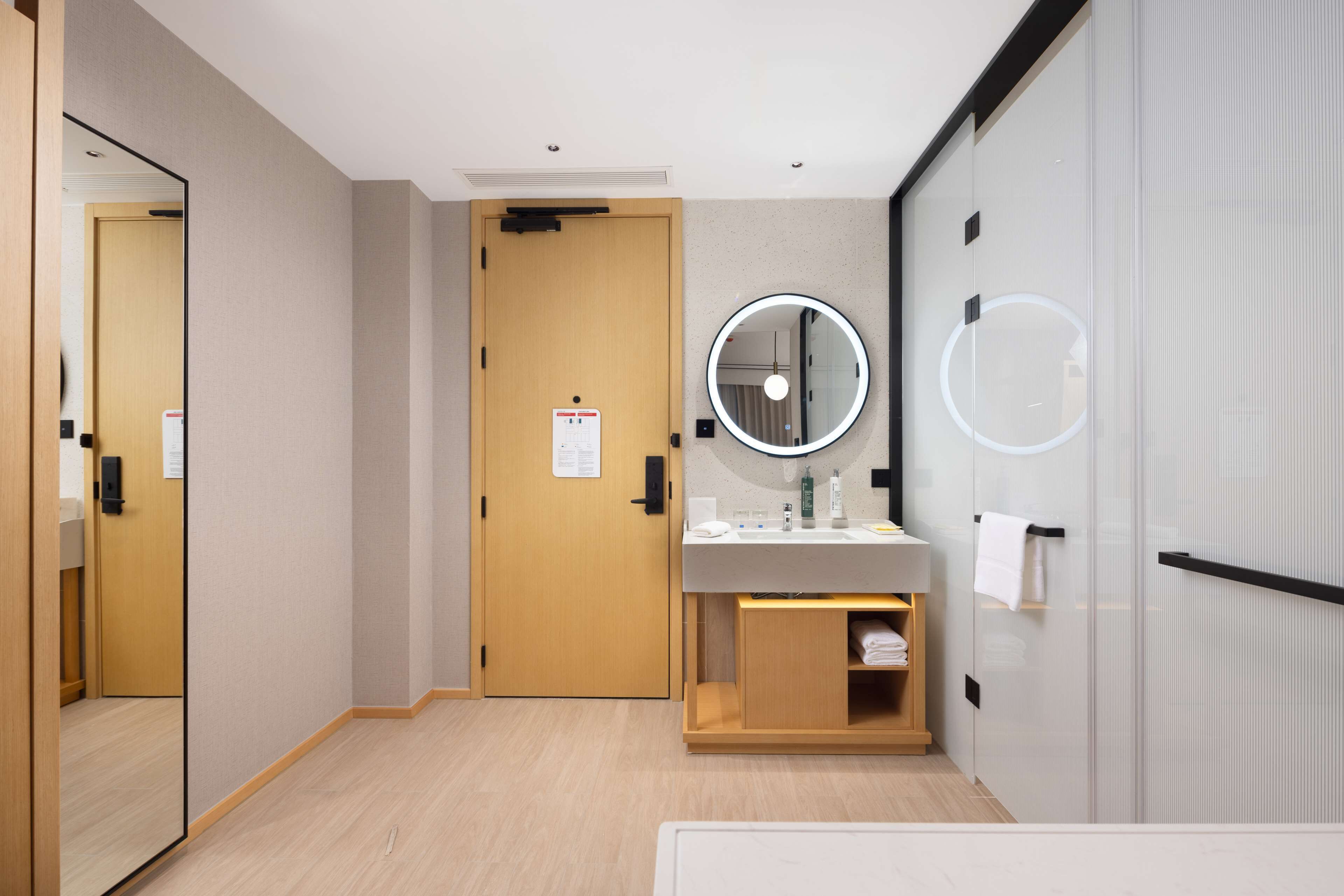 A hotel room with a wooden door, a round mirror, a sink, and a towel rack.