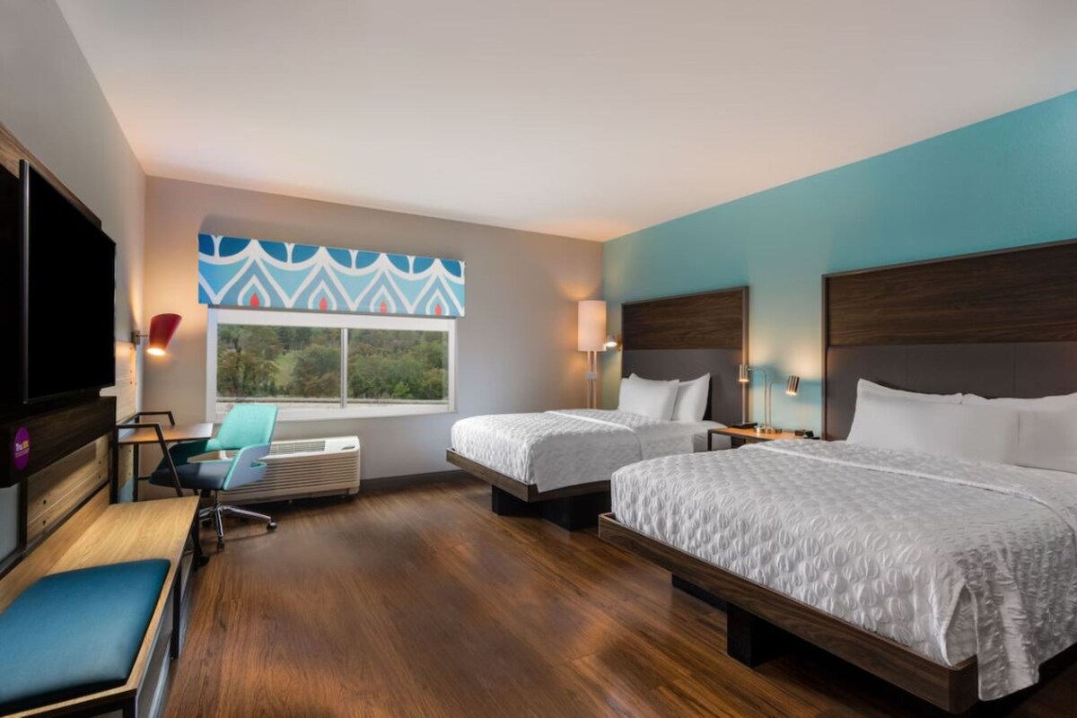A hotel room with two beds, a desk, a chair, a TV, and a window with a view of greenery.