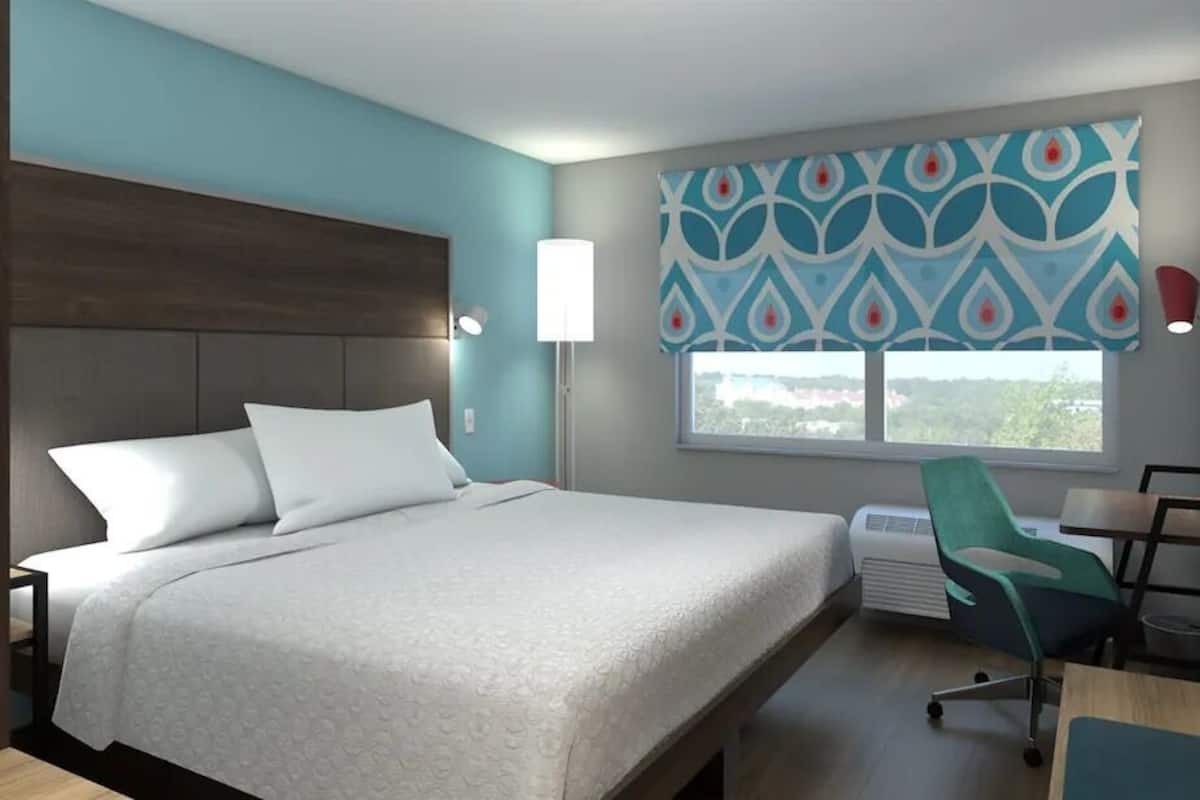 A hotel room with a large bed, a desk, a chair, a window with a view, and a flat-screen TV mounted on the wall.