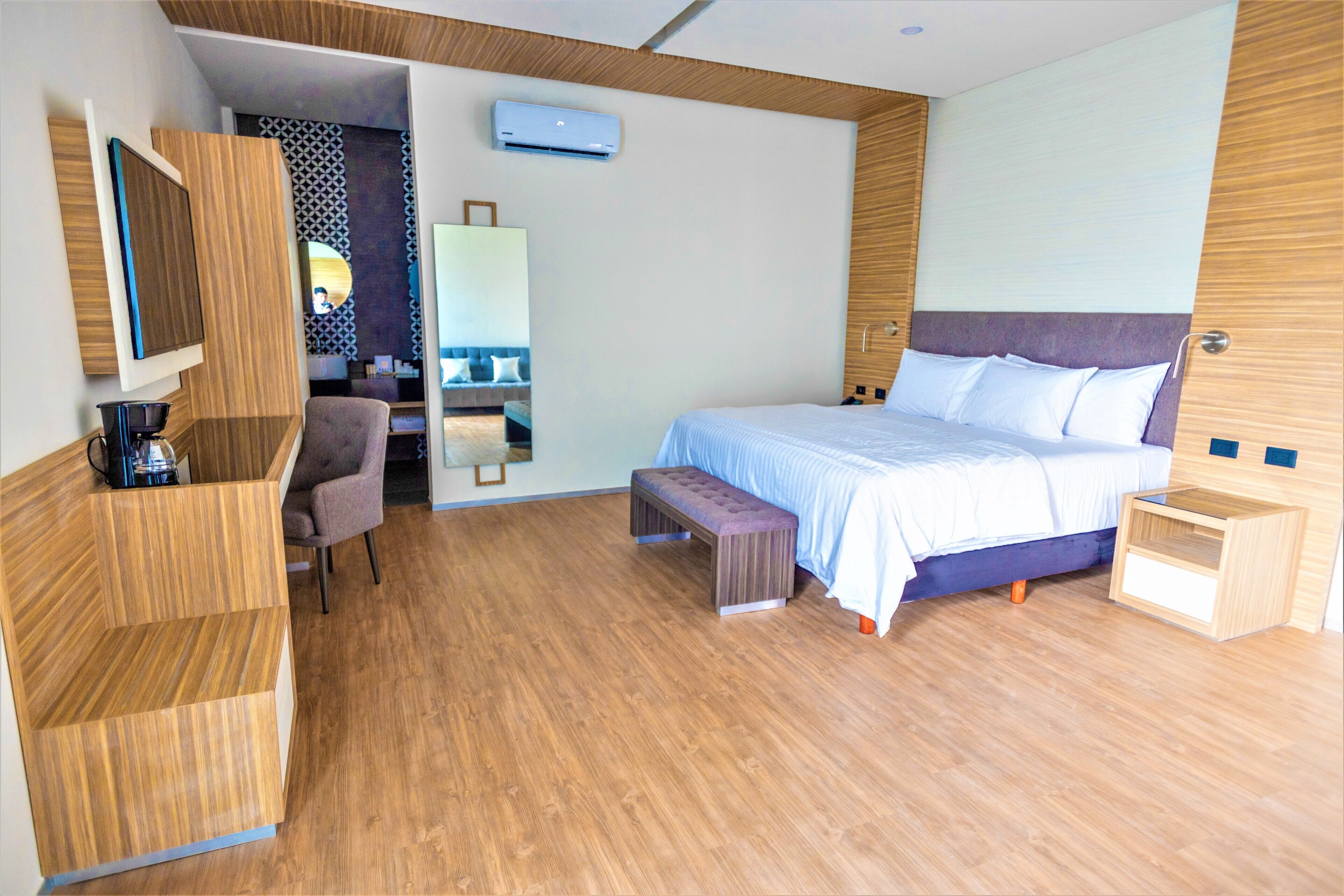 A modern hotel room with a wooden floor, a bed with white bedding, a wooden desk with a chair, a small bench, and a mirror.