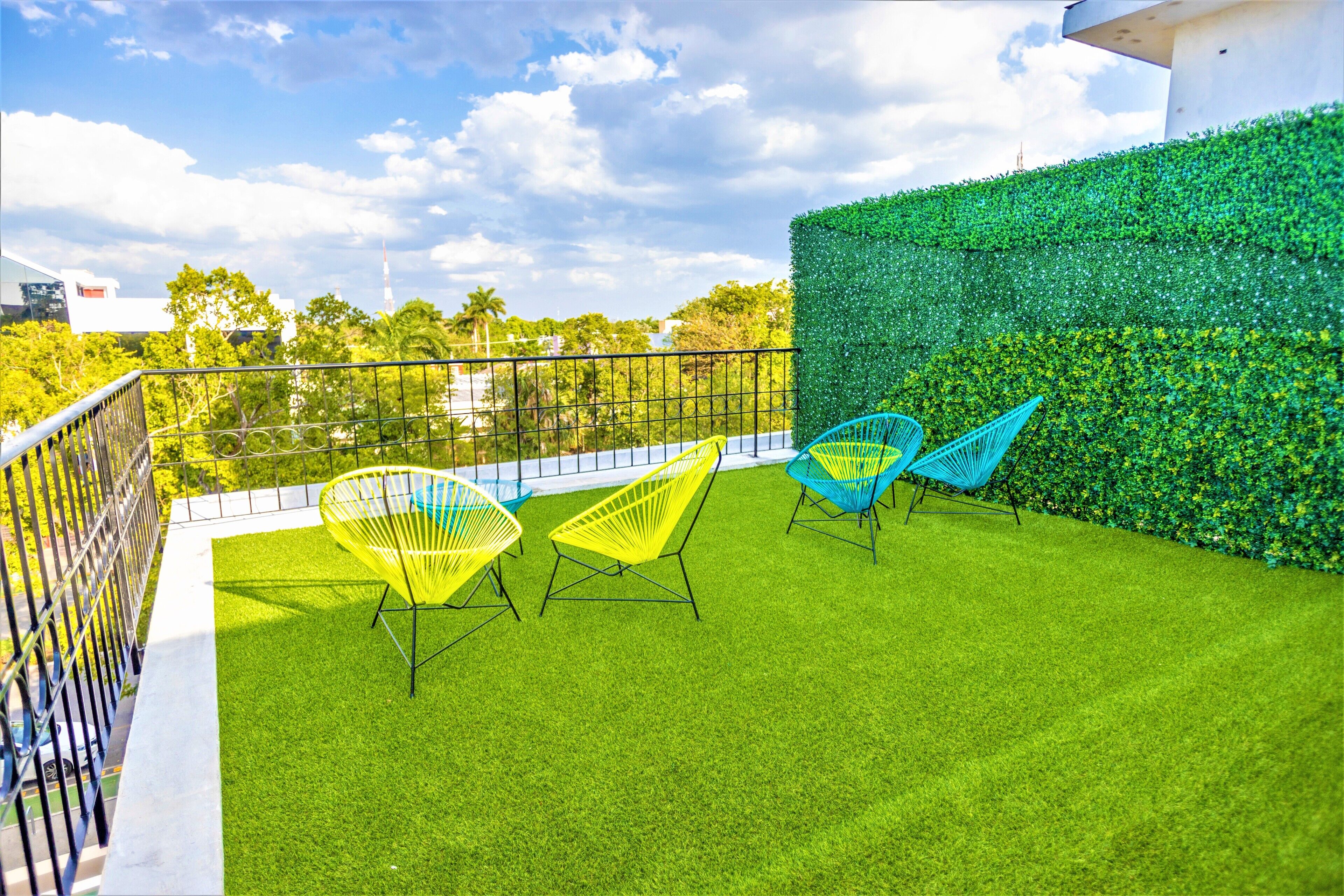 Una terraza en la azotea con césped artificial, tres sillas amarillo brillante y una azul, y un muro de seto verde.