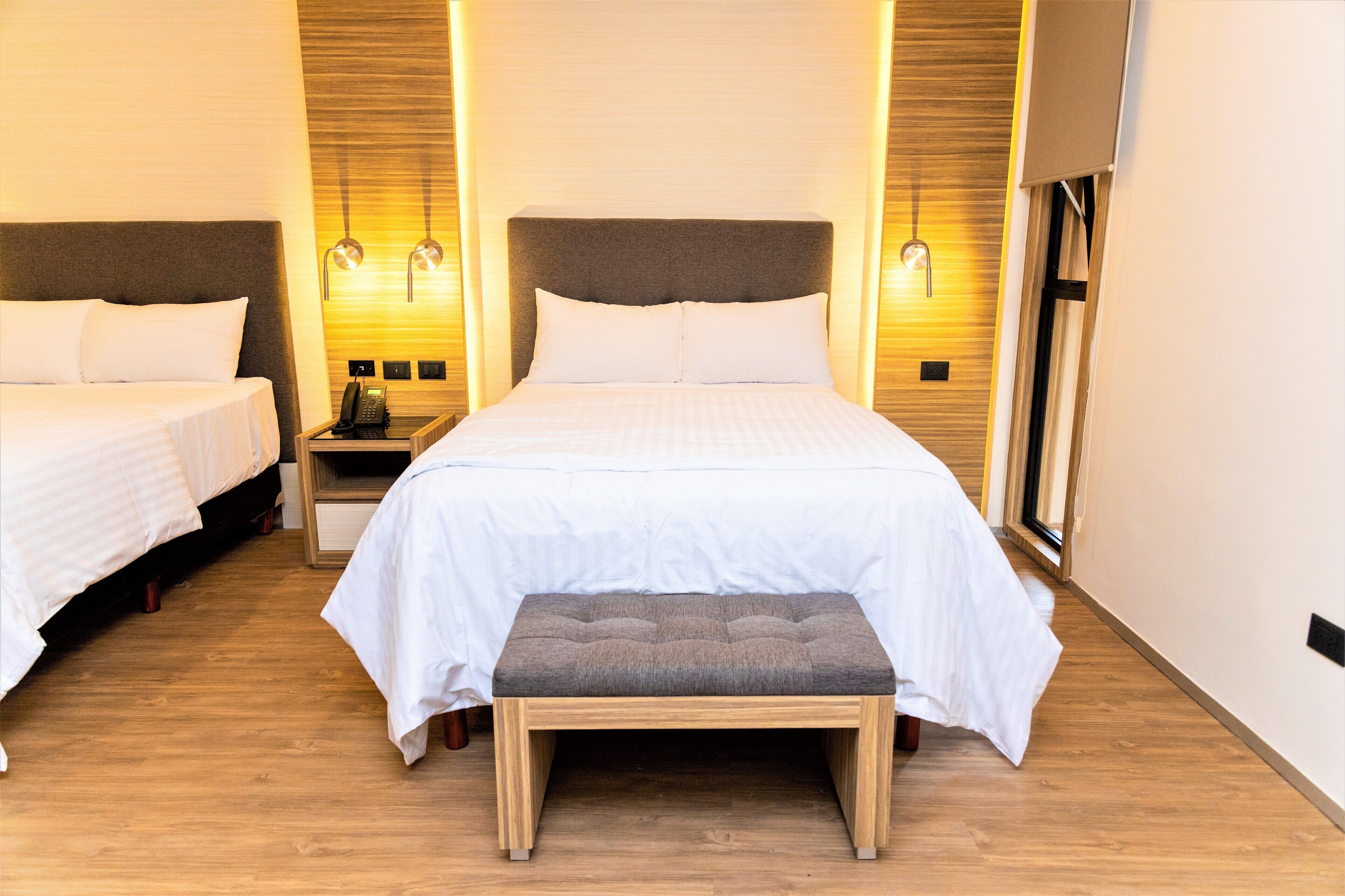 Habitación de hotel con dos camas, un banco, mesitas de noche y lámparas de pared.
