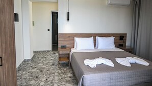 Una habitación de hotel con una cama, una mesita de noche, un cabecero de madera, un aire acondicionado instalado en la pared y una puerta que da a otra habitación.