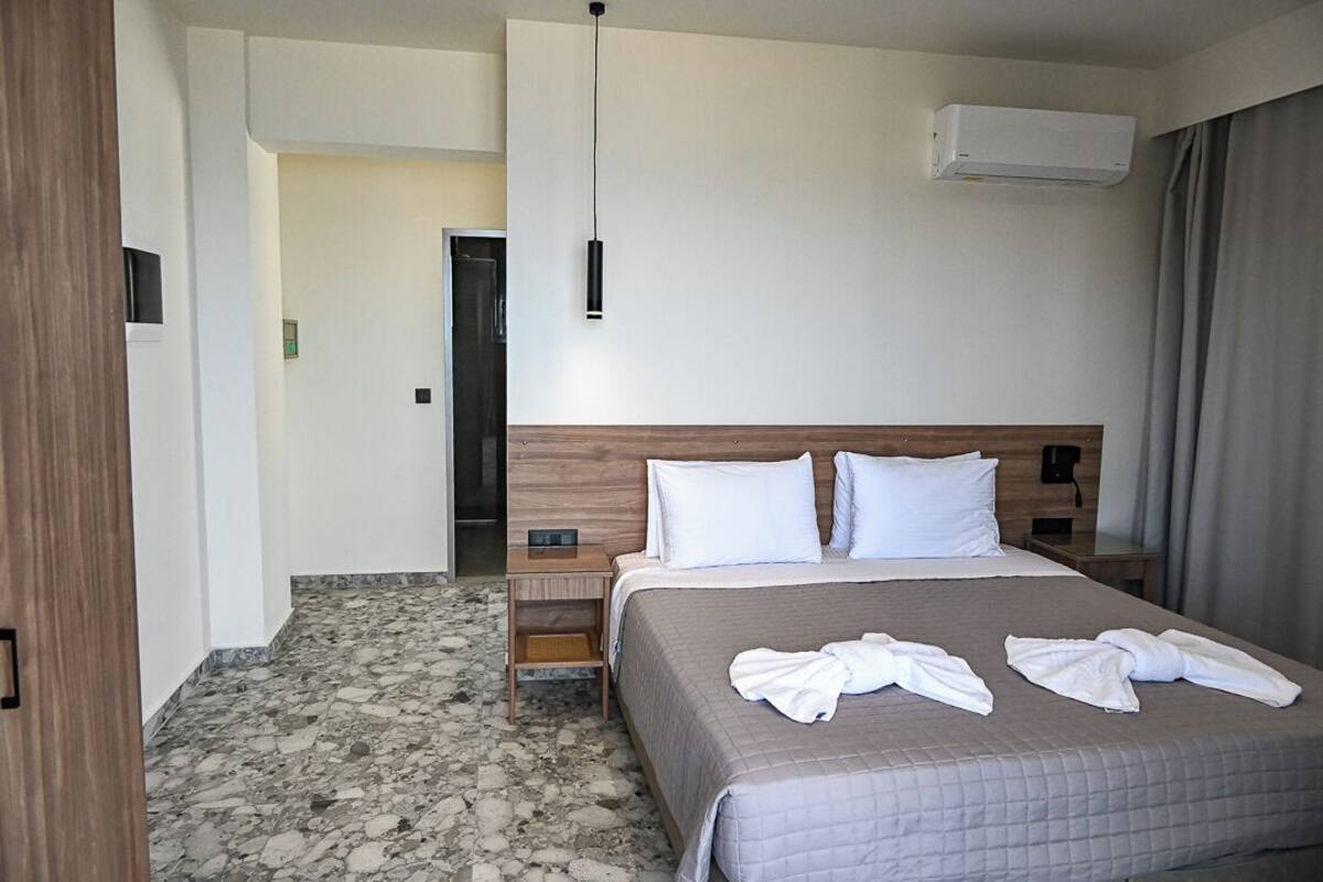 A hotel room with a bed, a nightstand, a wooden headboard, a wall-mounted air conditioner, and a door leading to another room.