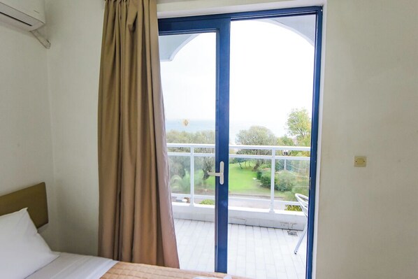A bedroom with a bed, a window with curtains, and a view of a balcony with greenery. - Amalia Sea Hotel (Rhodes)