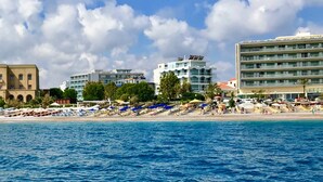 Un hotel costero con varios pisos, una playa con tumbonas y sombrillas, y un cielo azul despejado.