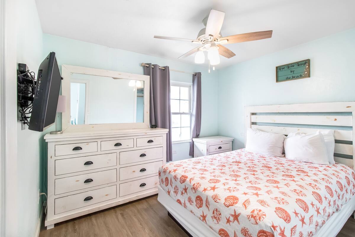 A bedroom with a bed, a dresser, a mirror, a ceiling fan, and a window with curtains.