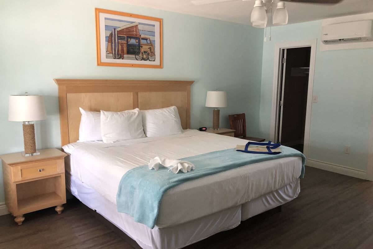 A neatly made bed with white linens, a blue throw blanket, and white towels. There are two bedside tables with lamps, a wooden headboard, and a framed picture on the wall.