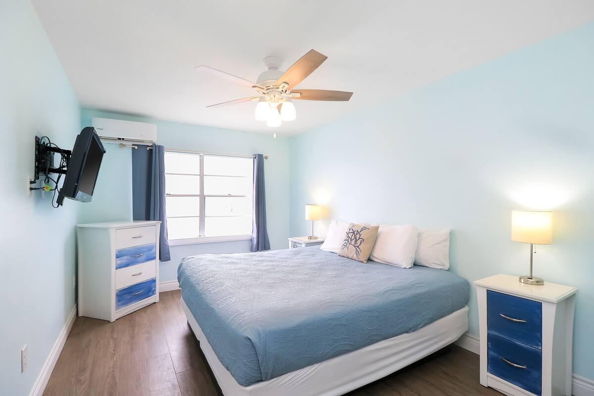 A bedroom with a bed, a ceiling fan, a dresser, a nightstand, and a lamp.
