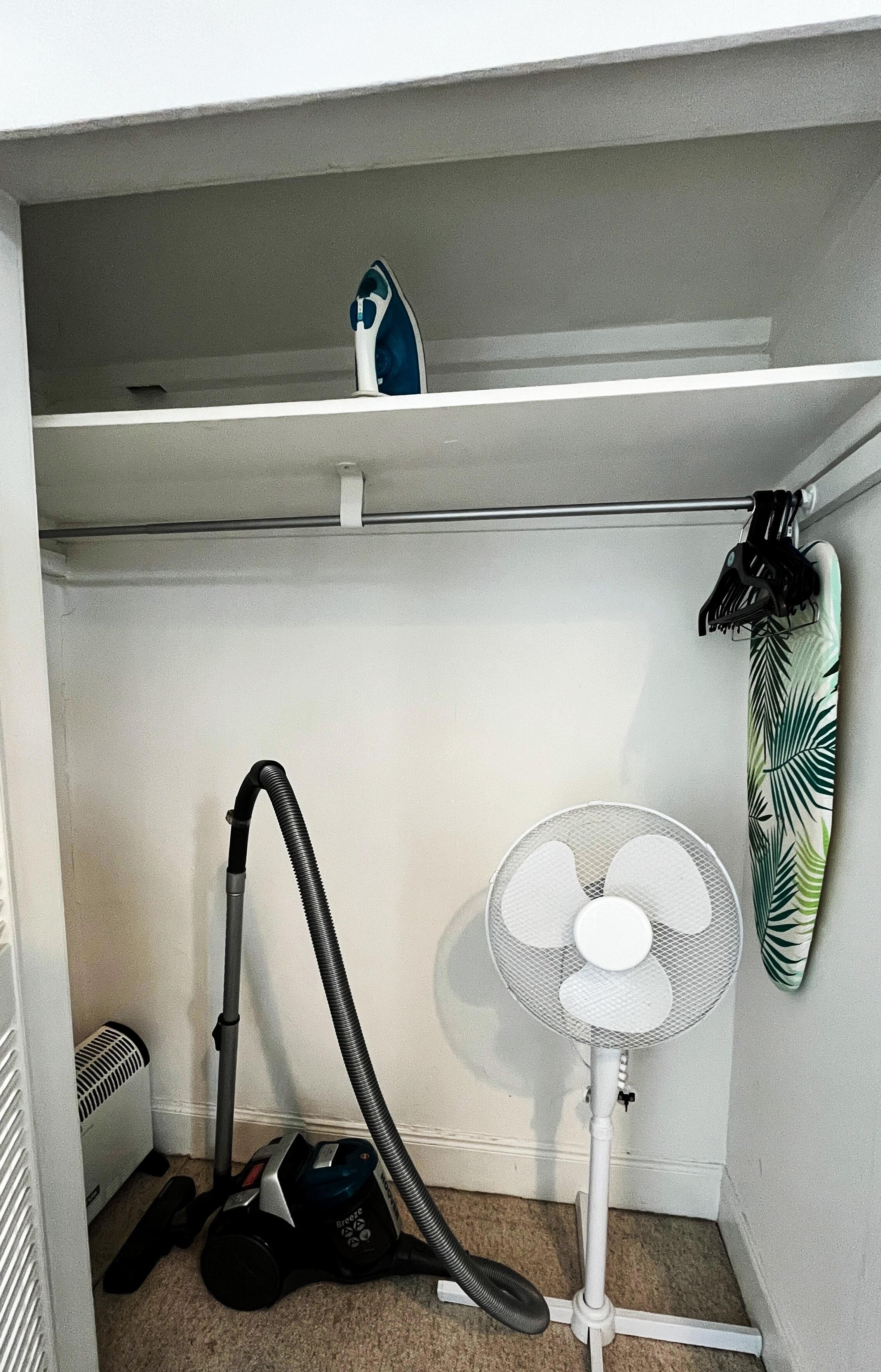 Een hoek van een kamer met een stofzuiger, een staande ventilator en een handdoekrek.
