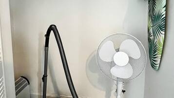 Een hoek van een kamer met een stofzuiger, een staande ventilator en een handdoekrek.