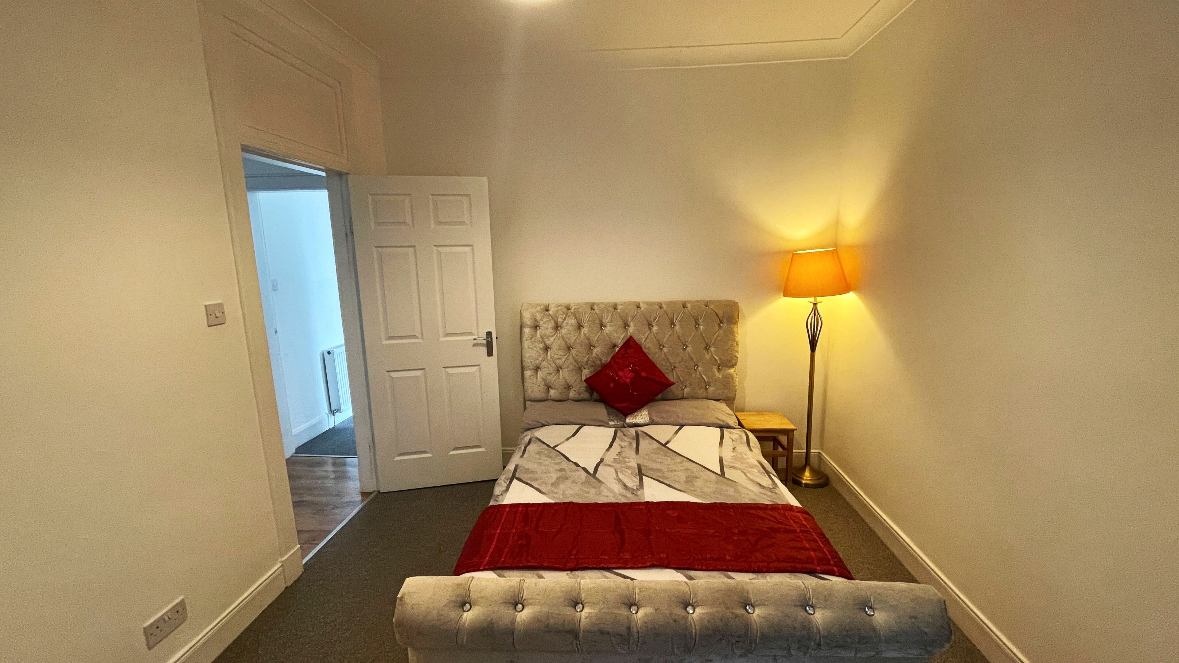 A bedroom with a bed, a bedside lamp, a bench, and a door leading to another room.