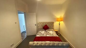 A bedroom with a bed, a bedside lamp, a bench, and a door leading to another room.