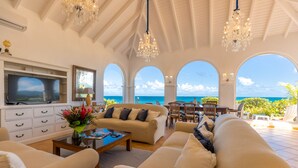 Un salón amplio con un televisor grande, asientos cómodos y vistas al océano a través de ventanas arqueadas.