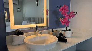Une salle de bain avec un lavabo blanc, un miroir, une serviette, une orchidée en pot et un sèche-cheveux noir.